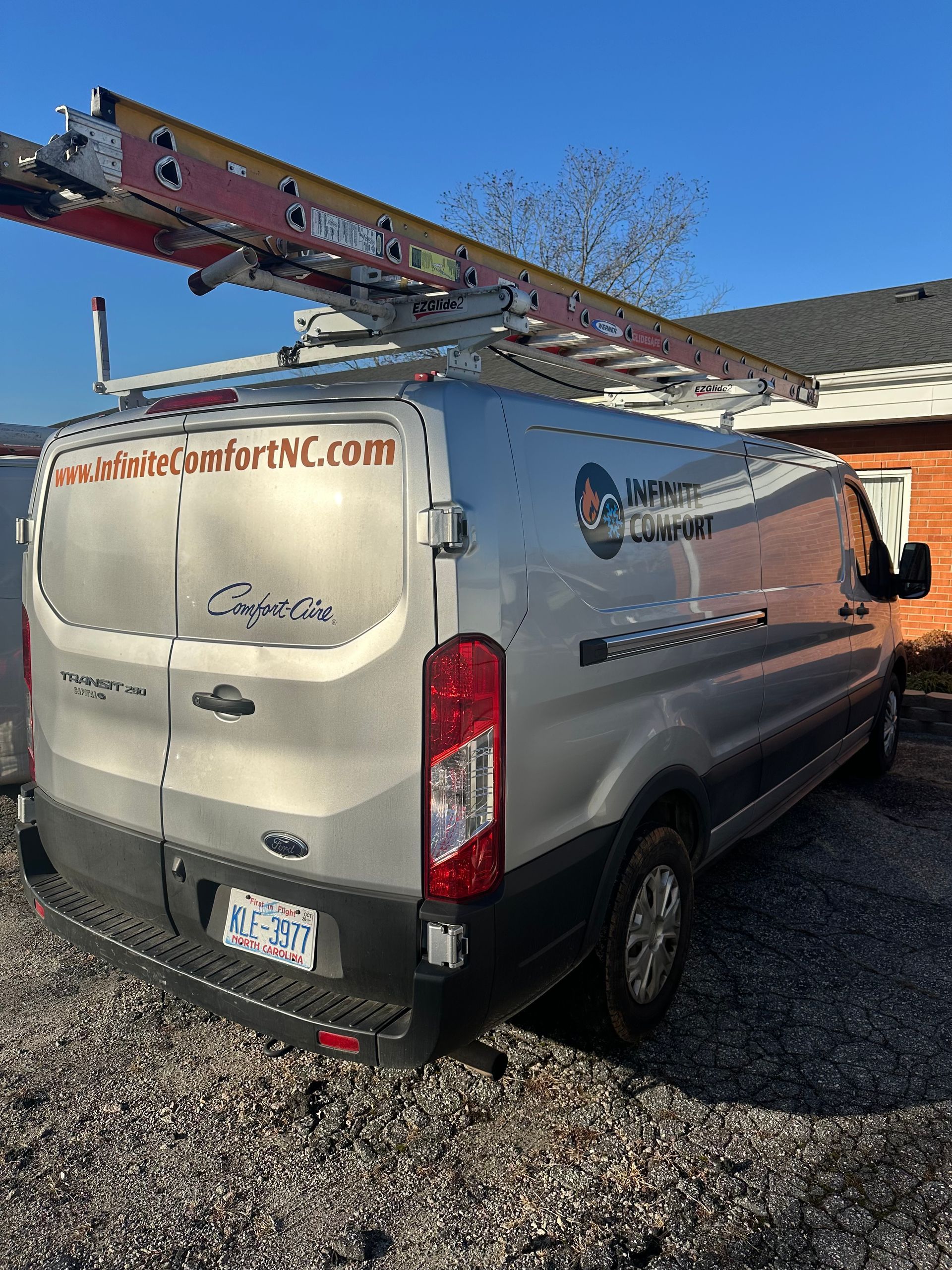 A silver Ford commercial cargo van with a ladder on its roof rack, parked on a gravel surface under a clear blue sky.
