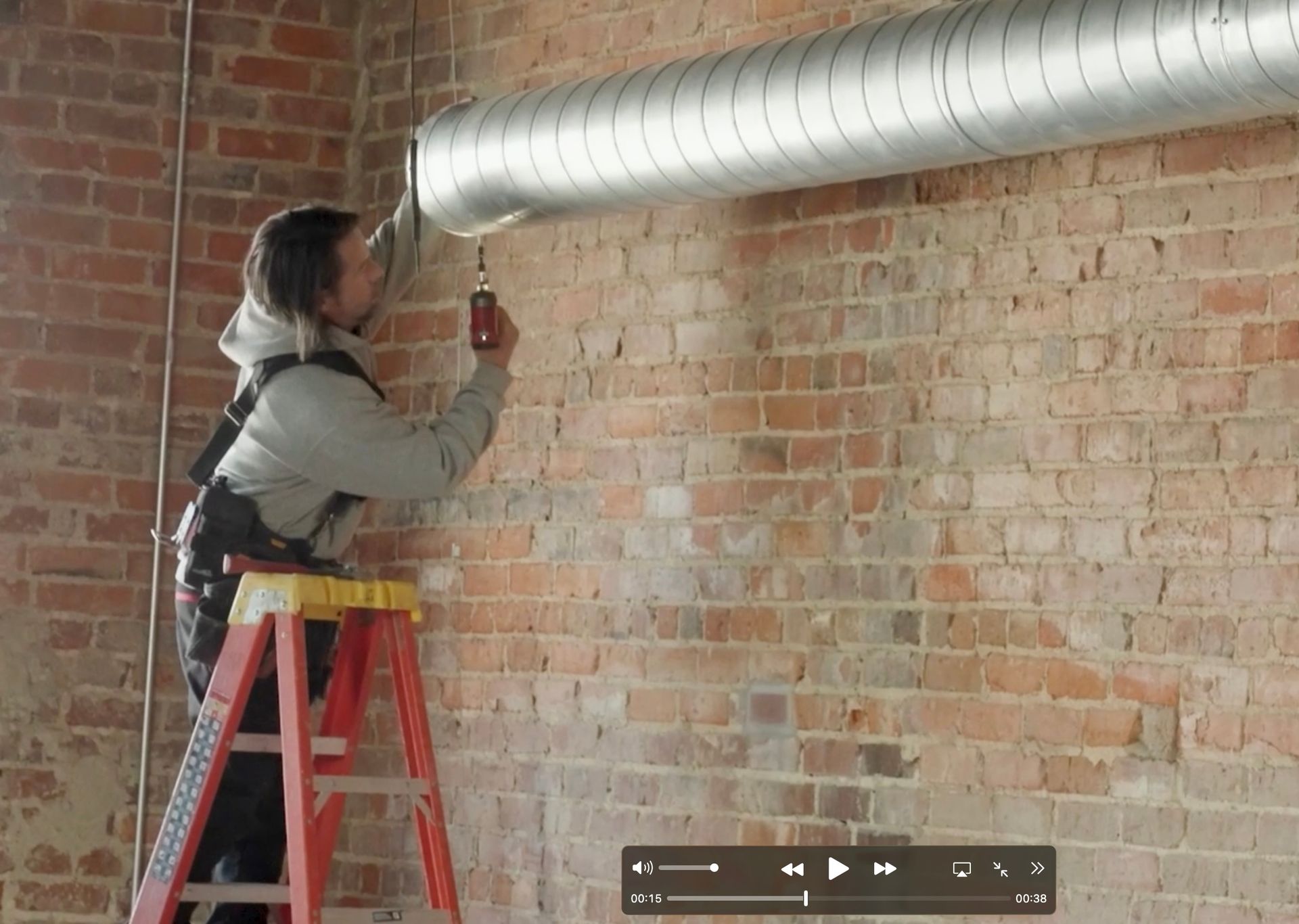 A person on a red step ladder uses a cordless drill to install a silver metal HVAC duct onto an exposed brick wall.