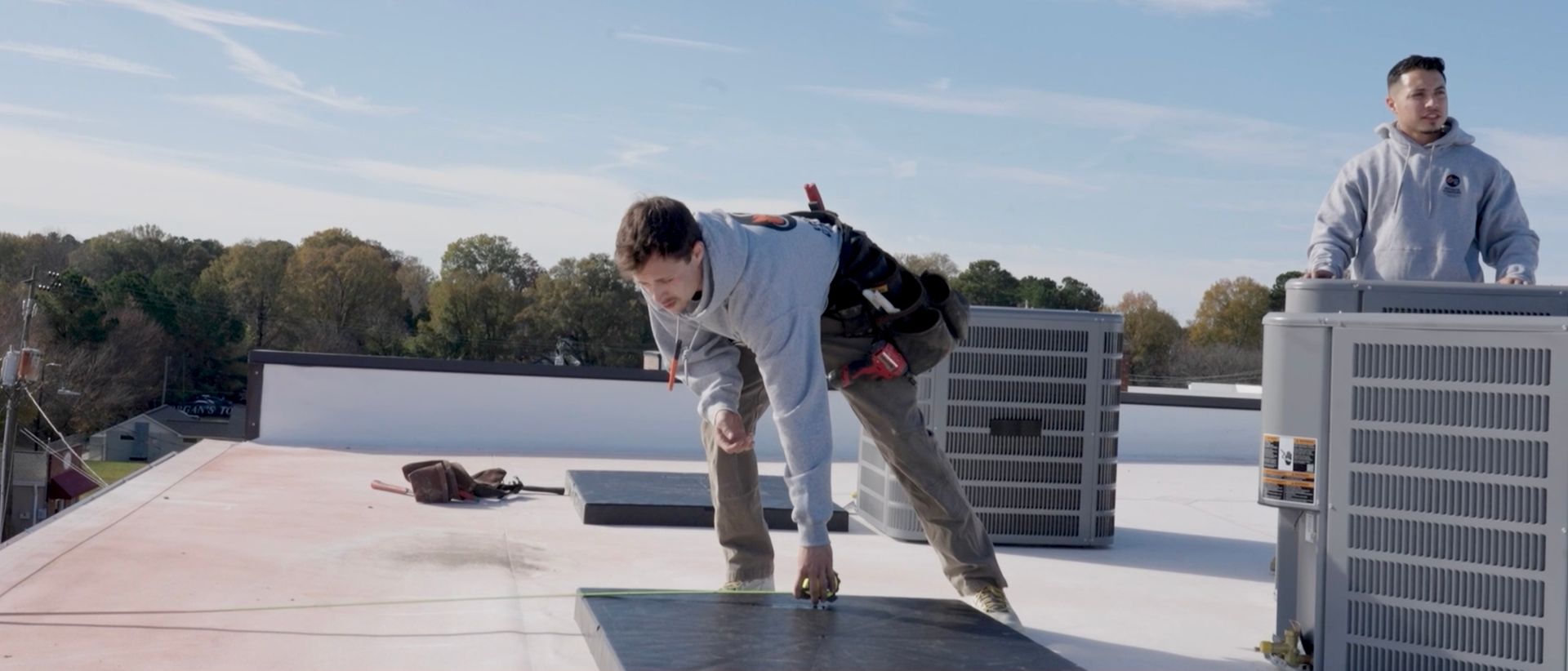 Two workers in hoodies install equipment on a flat building roof against a clear blue sky.