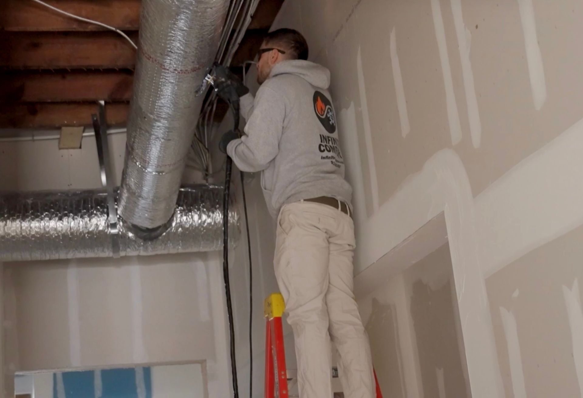 A worker wearing a gray hoodie stands on a ladder, working on ventilation ducts attached to exposed wooden ceiling joists.