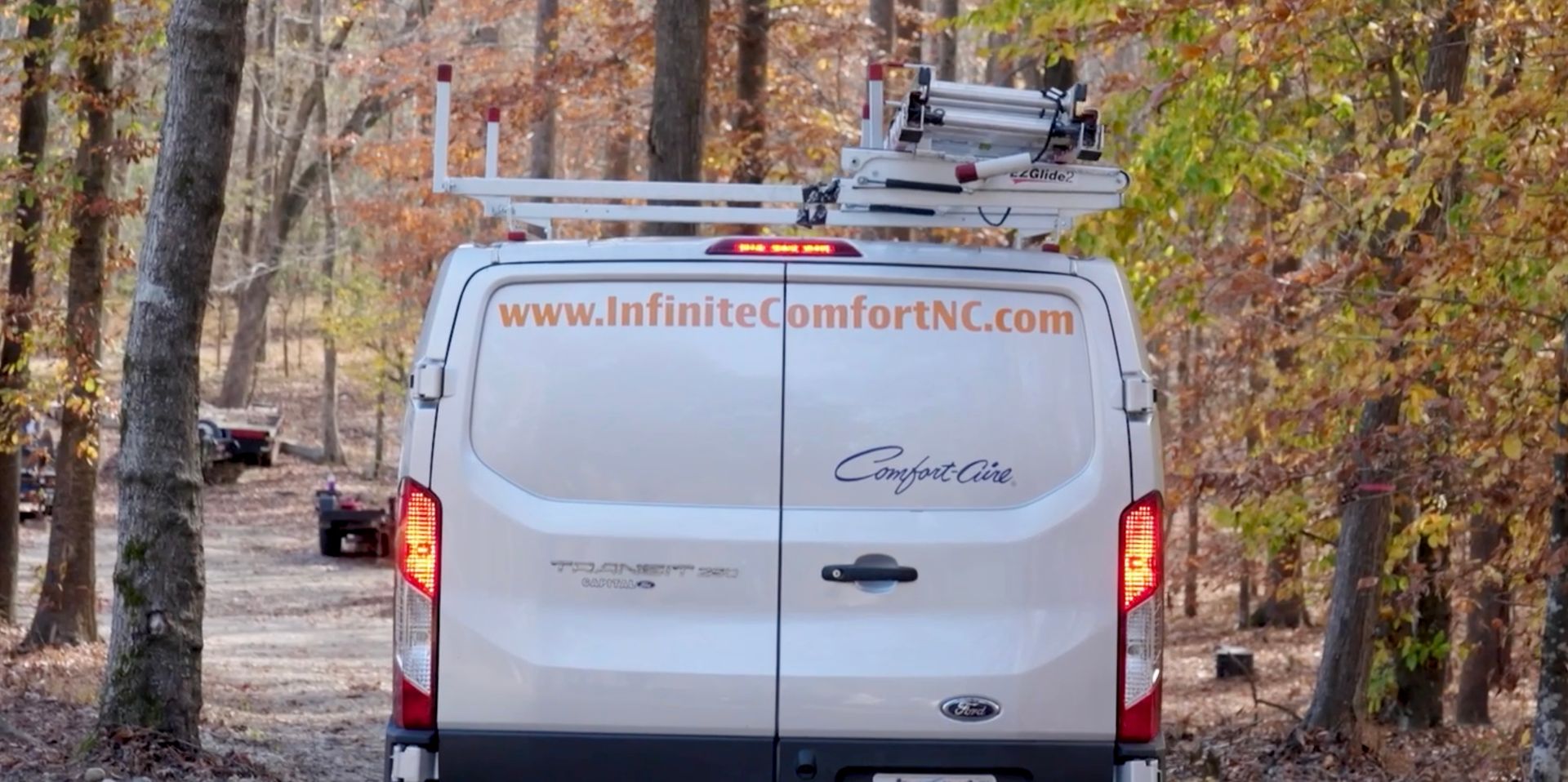 A white work van with a ladder rack parked on a dirt path surrounded by trees.