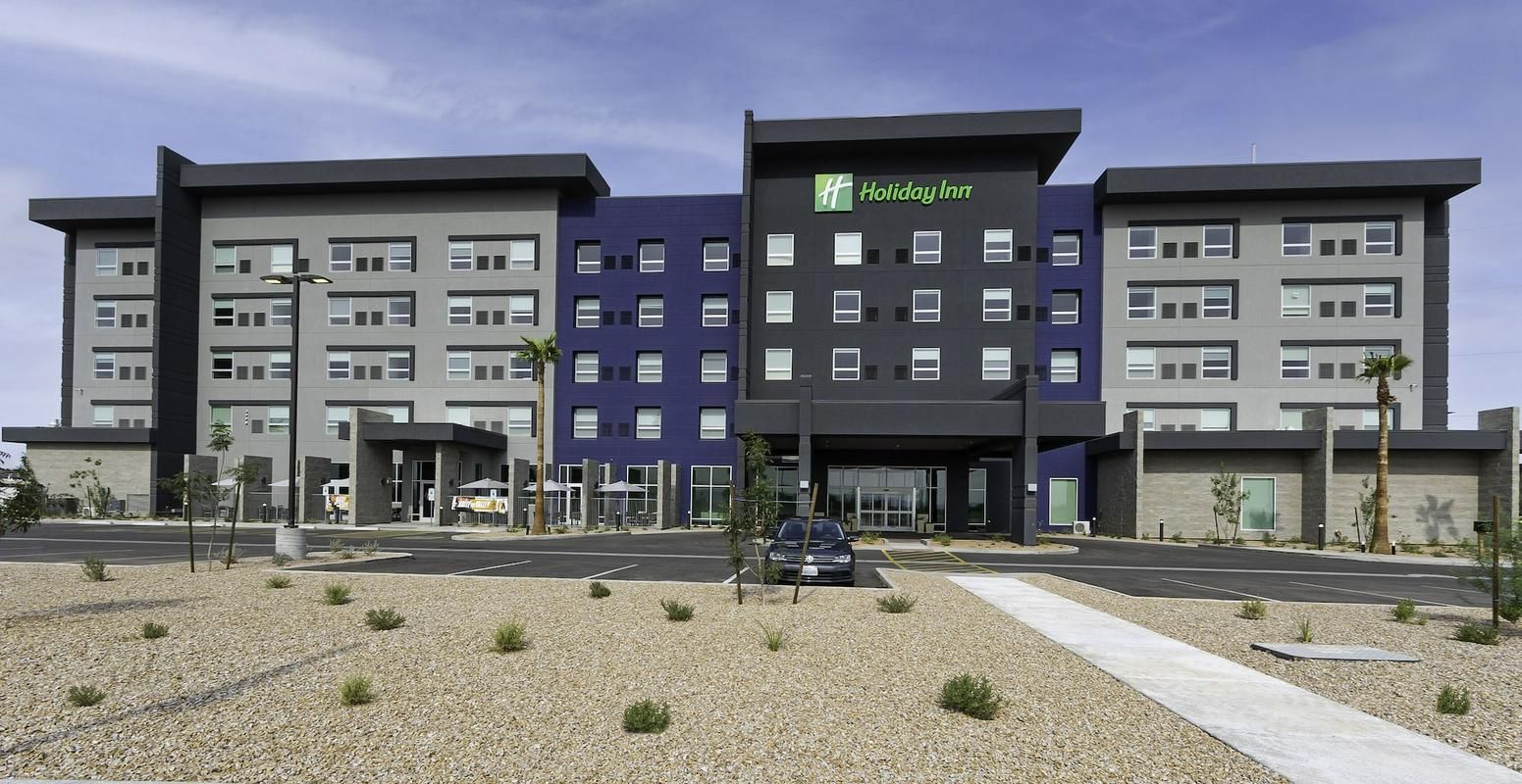 A four-story Holiday Inn hotel with grey, blue, and black exterior walls, surrounded by a gravel landscape and a parking lot.