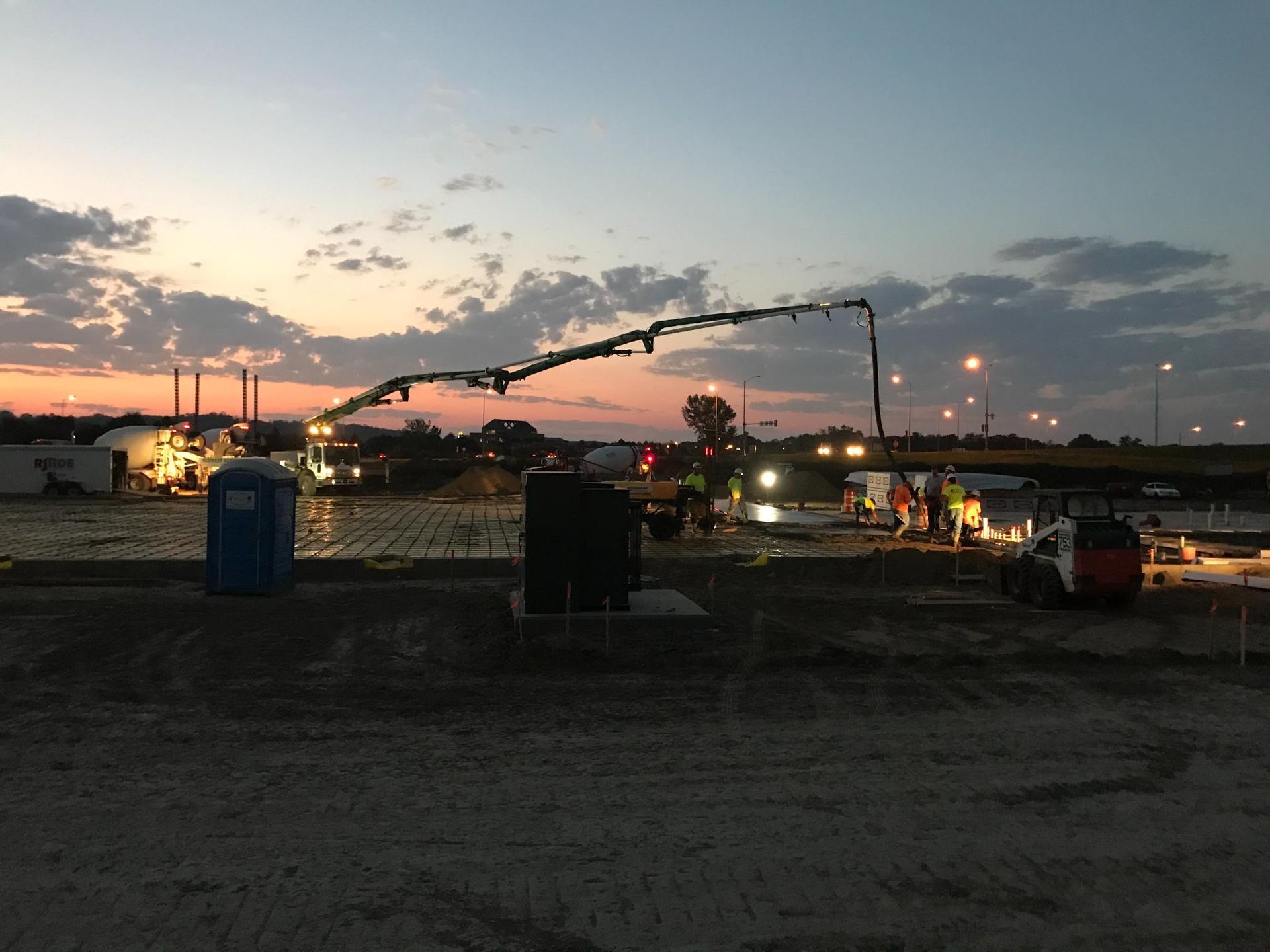 Dusk over a construction site with trucks, lights, and a blue portable toilet under a cloudy sky
