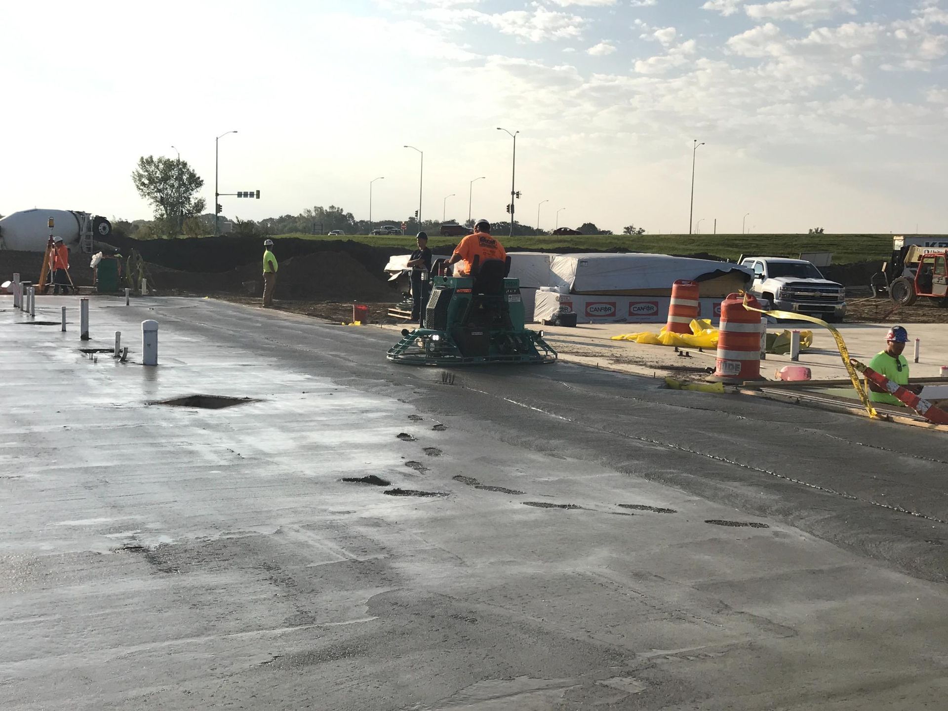Roadwork crew paving a wet road, with orange cones and barriers under a bright sky.