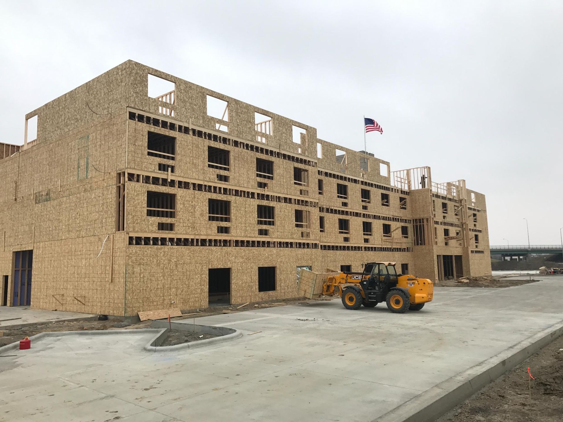 Partially built tan brick building with a yellow construction loader on a paved site under a cloudy sky
