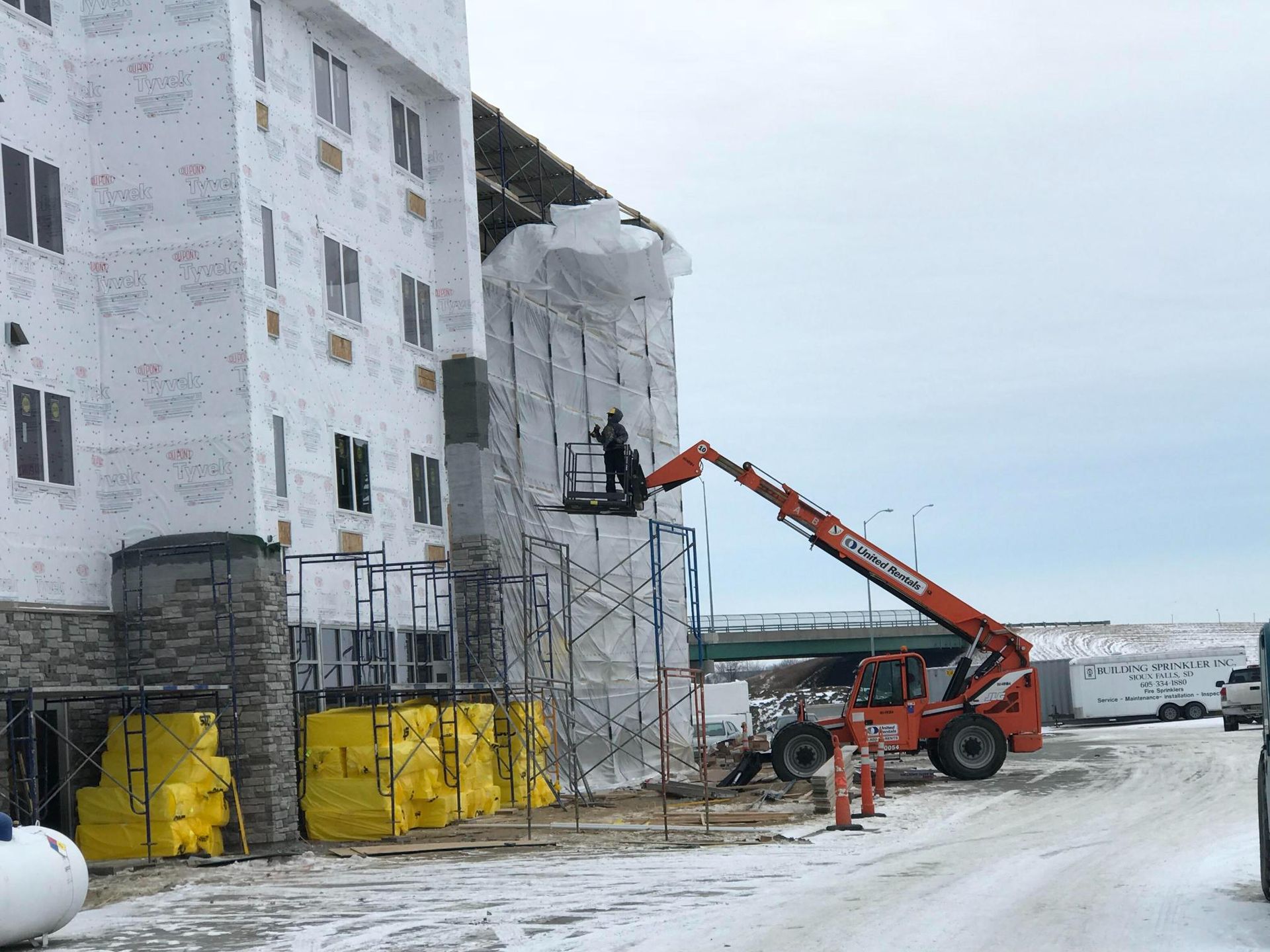 Construction workers in orange lift materials to a white building wrapped in scaffolding and yellow barriers.