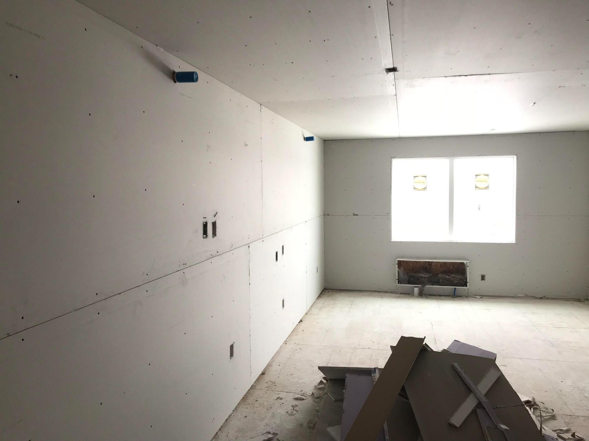 Unfinished white room with drywall walls, exposed ceiling, windows, and строитель materials piled on the floor.