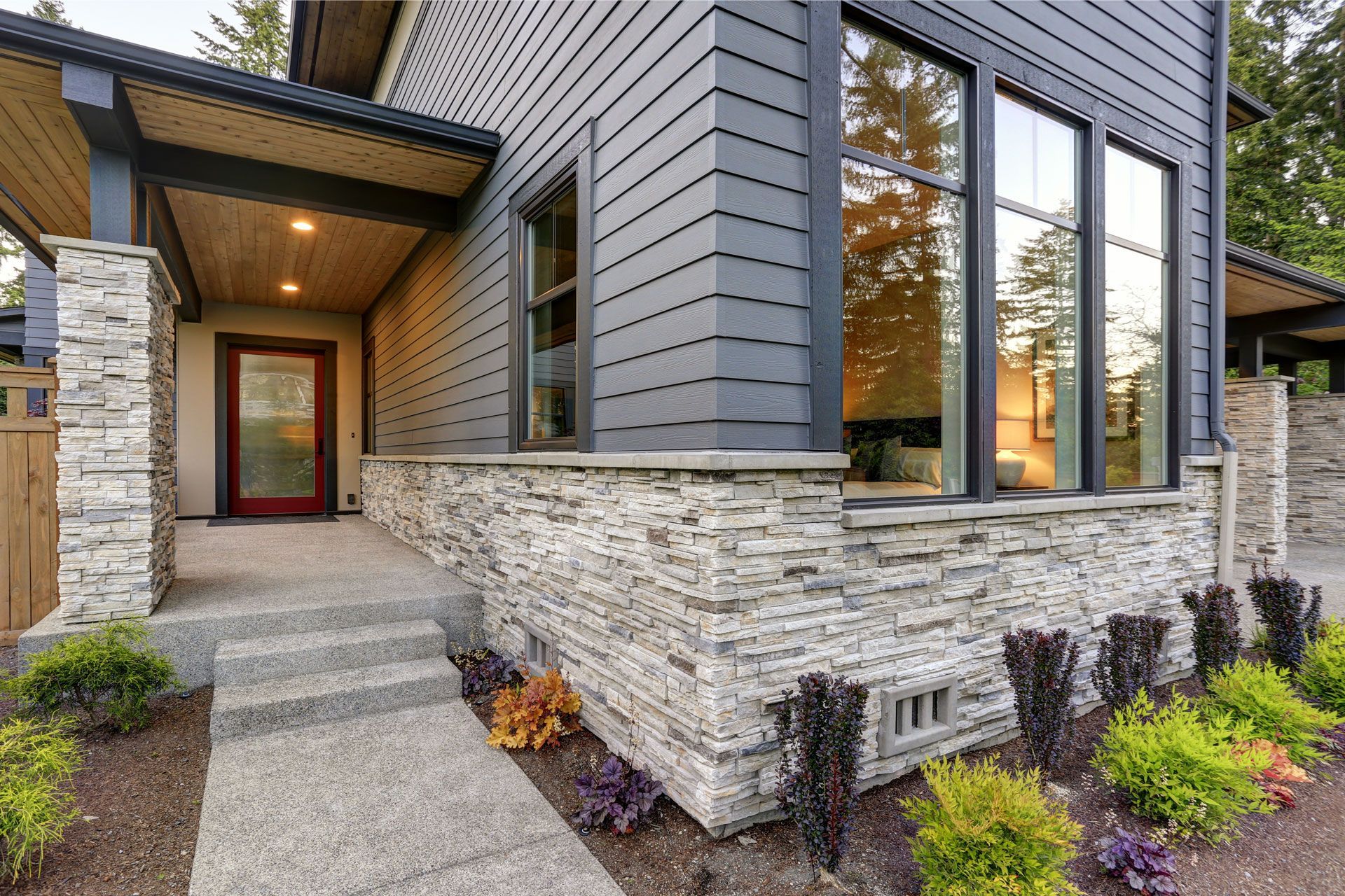 Modern house with gray siding, stone accents, large windows, and a red front door.