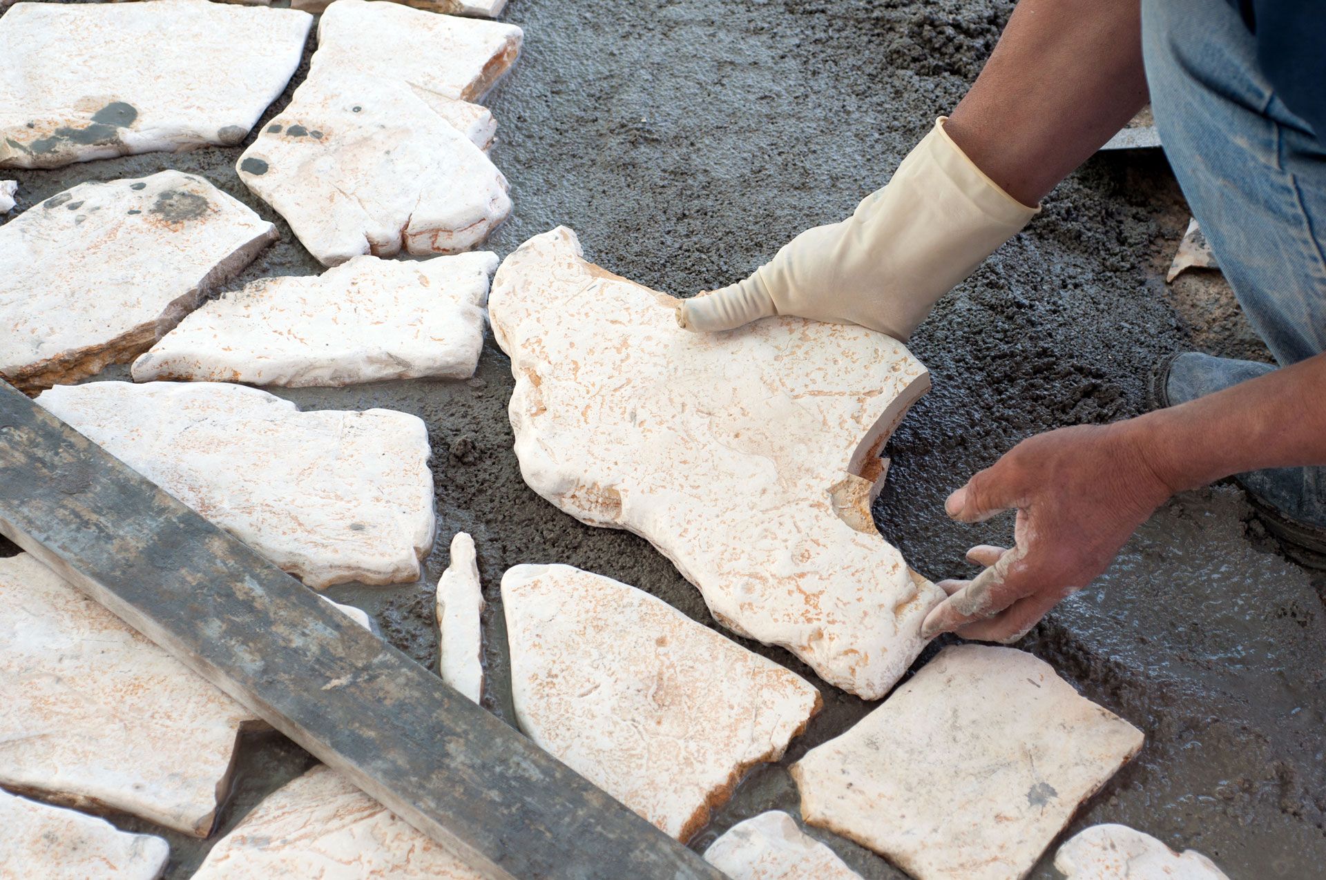 Person with gloves laying irregularly shaped stone pieces on a cement surface.