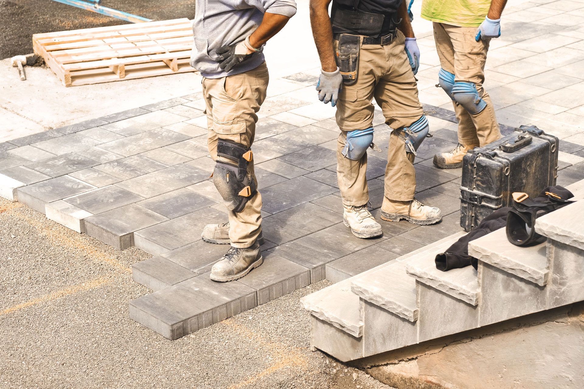 Three construction workers laying gray pavers outdoors; one on steps, the others standing near the work.