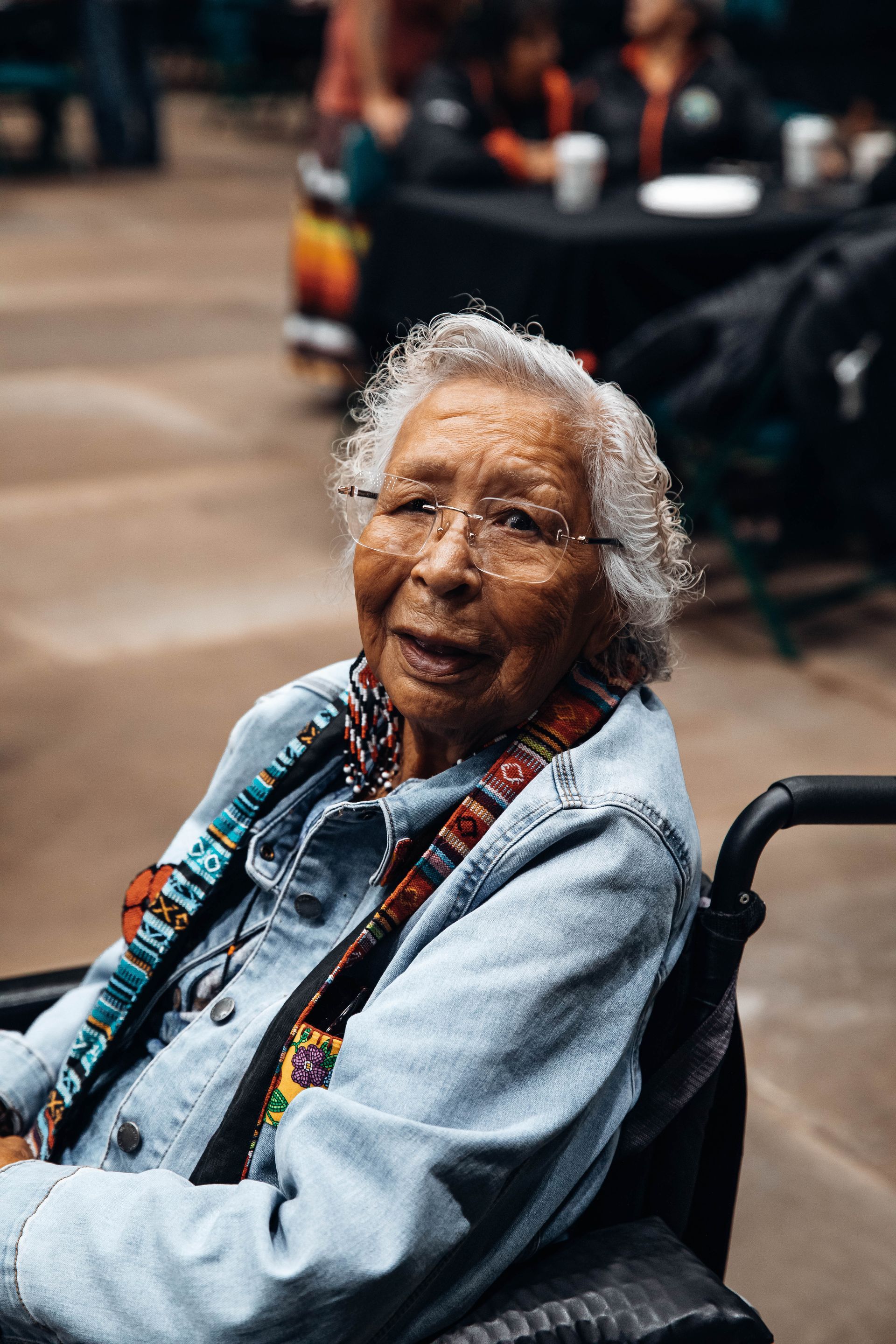 Elderly woman in a wheelchair wearing glasses and a denim jacket, smiling.