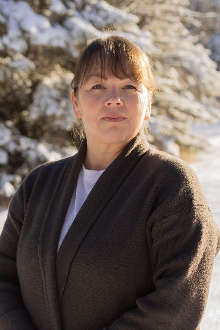 Woman wearing a brown cardigan stands outdoors with snow-covered trees in the background.