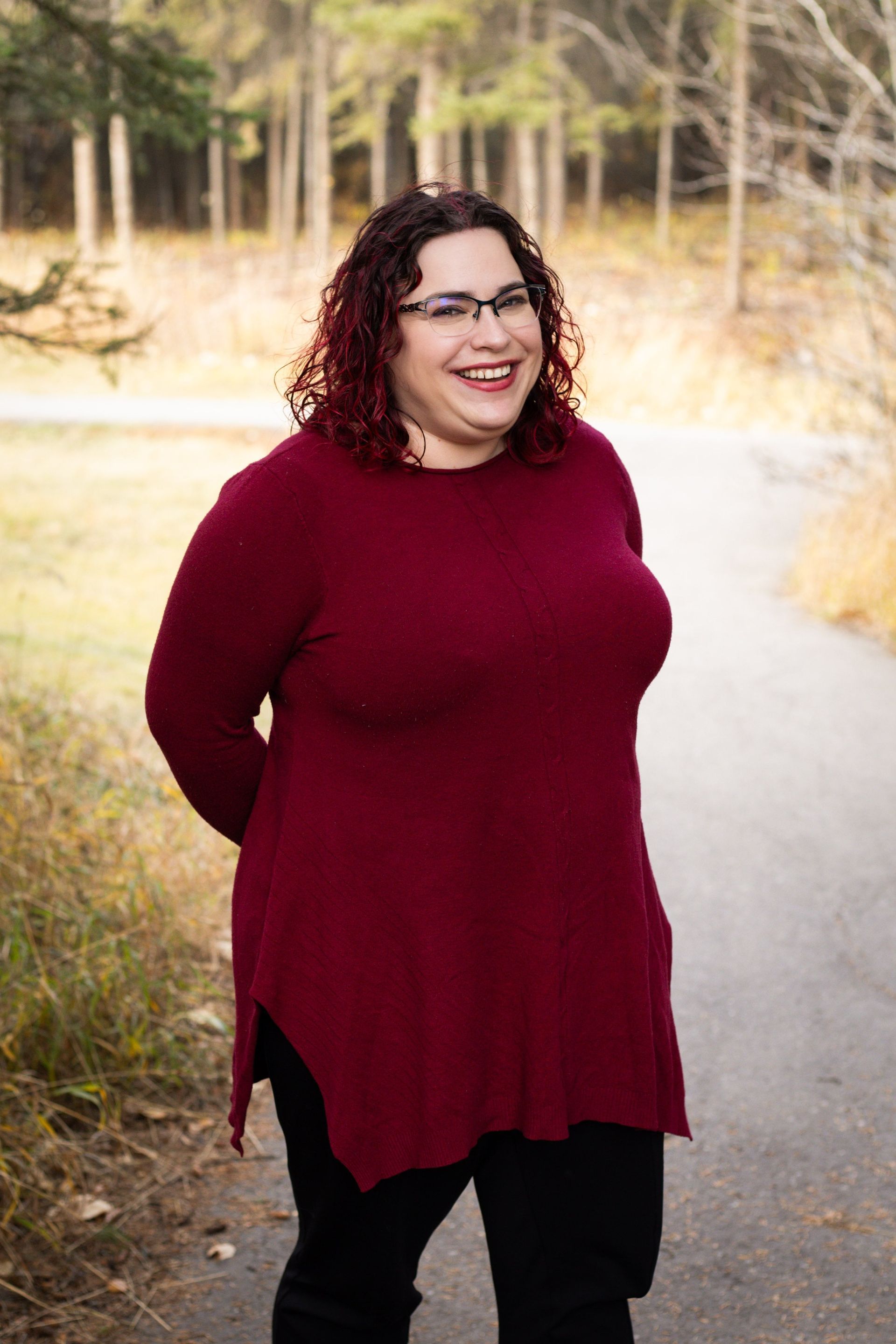Woman in burgundy sweater and glasses smiles on a path in a park.