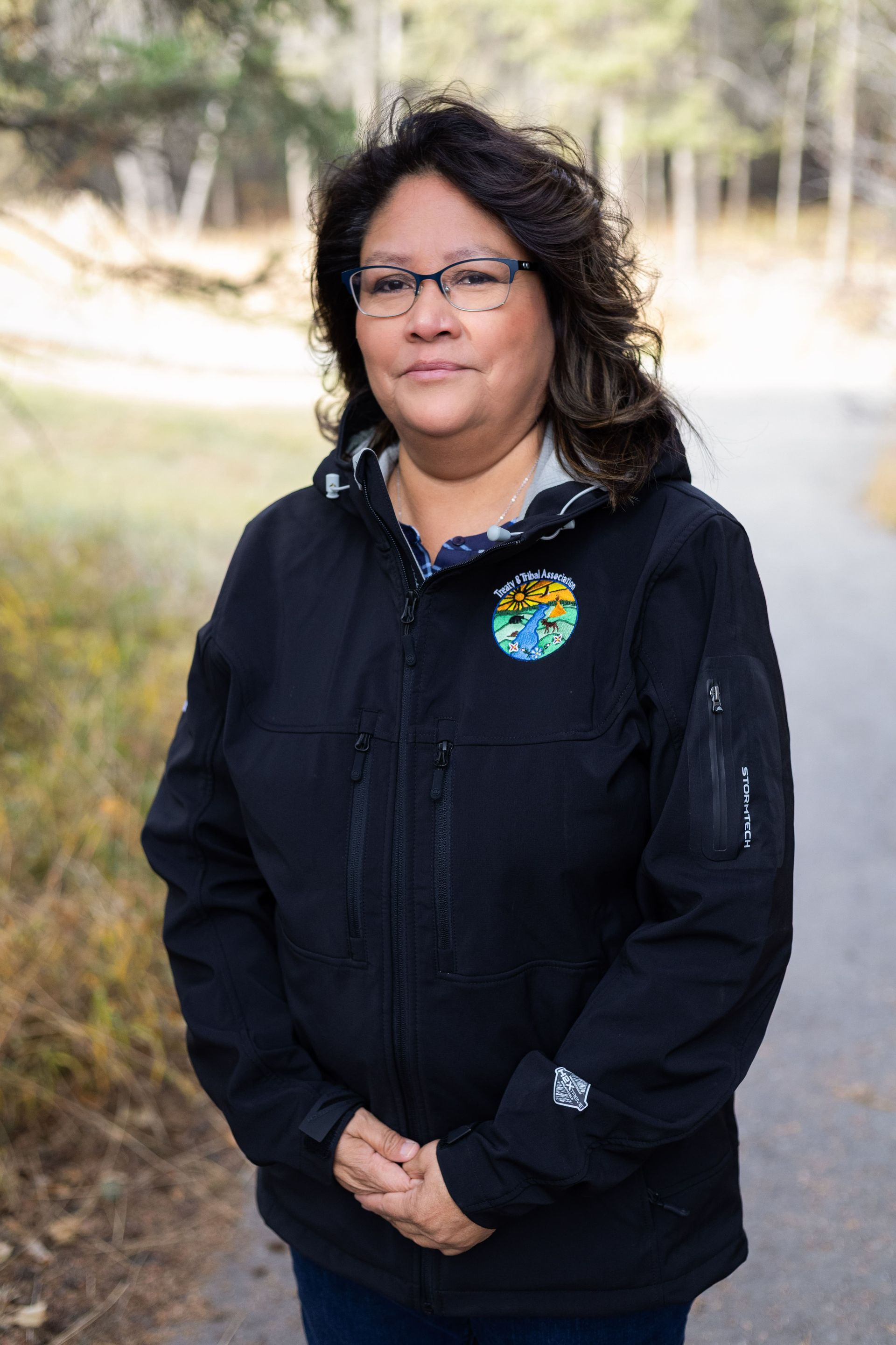 Woman in a black jacket stands on a path in a park, smiling slightly.