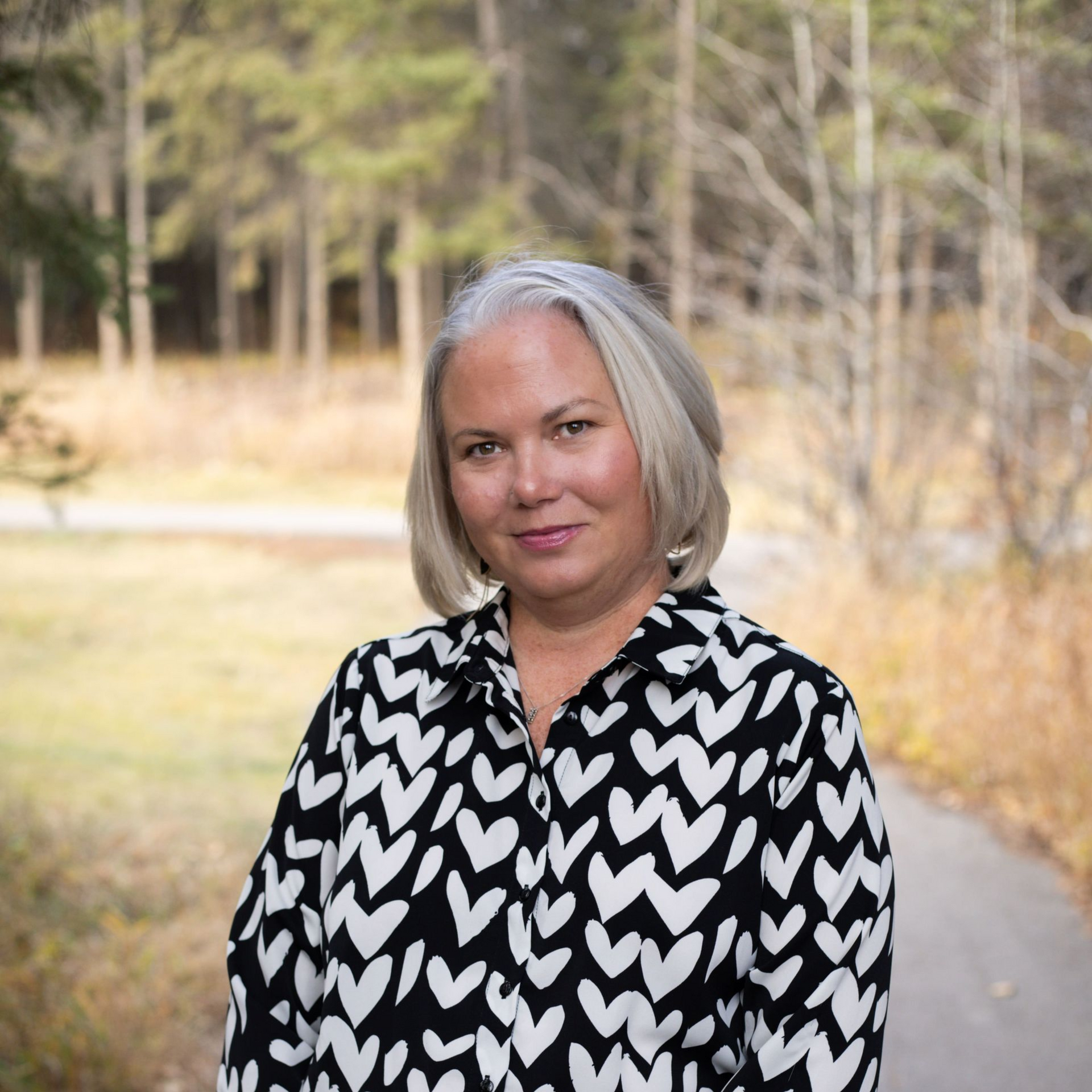 Woman with gray hair in black and white patterned shirt, standing outdoors in front of a forest.