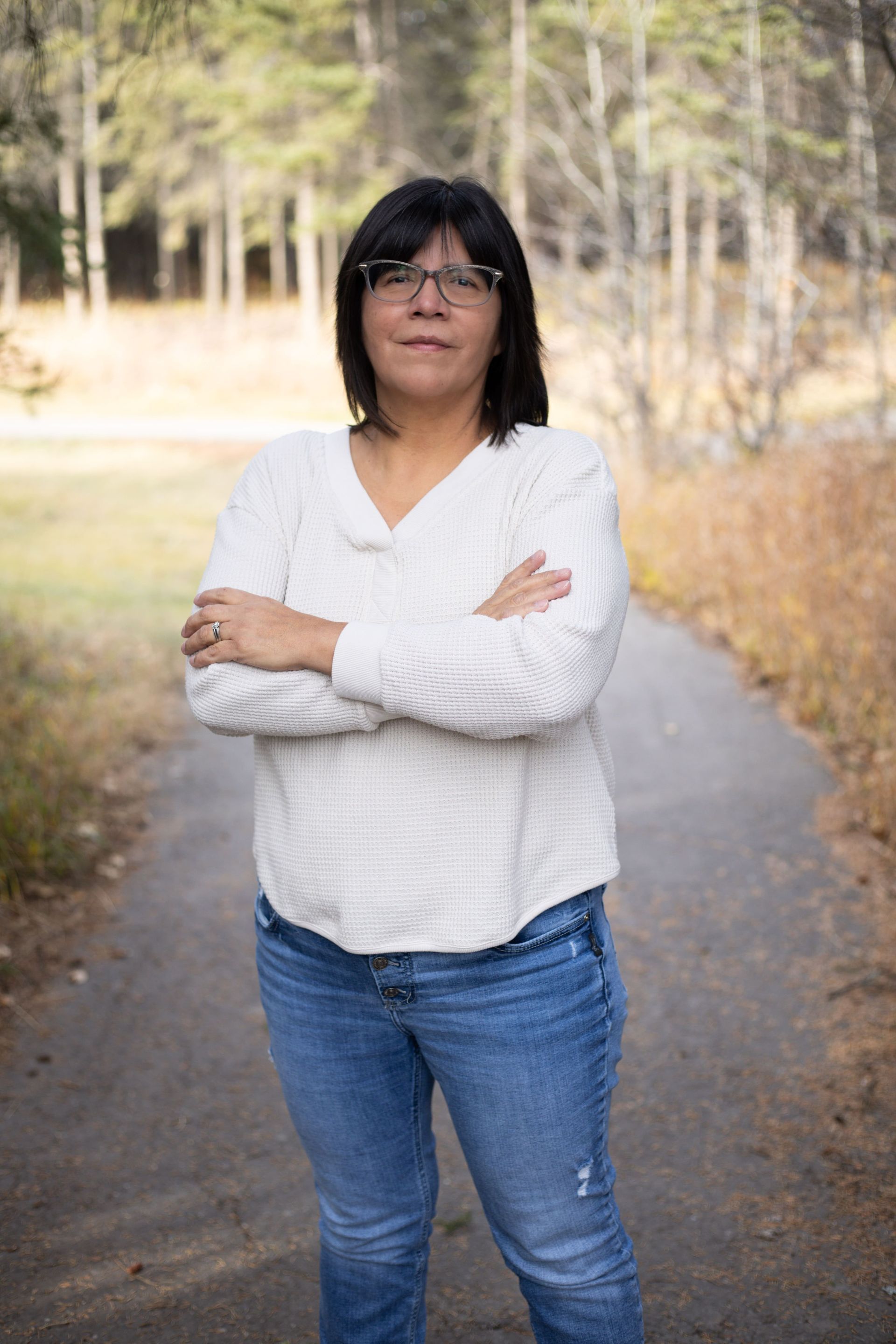 Woman with crossed arms, wearing glasses, standing on a path in a park, smiling.