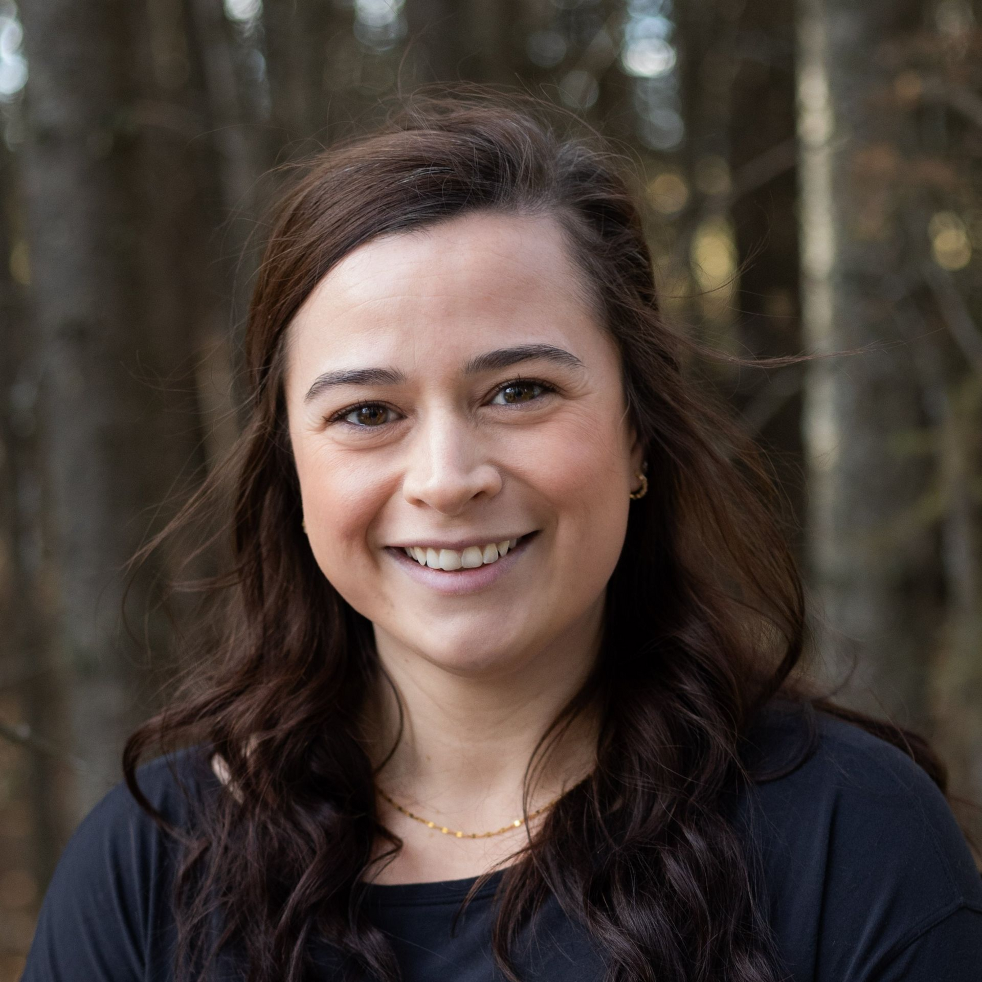 Woman smiling, brown hair, wearing a black shirt, outdoors with blurred trees in background.