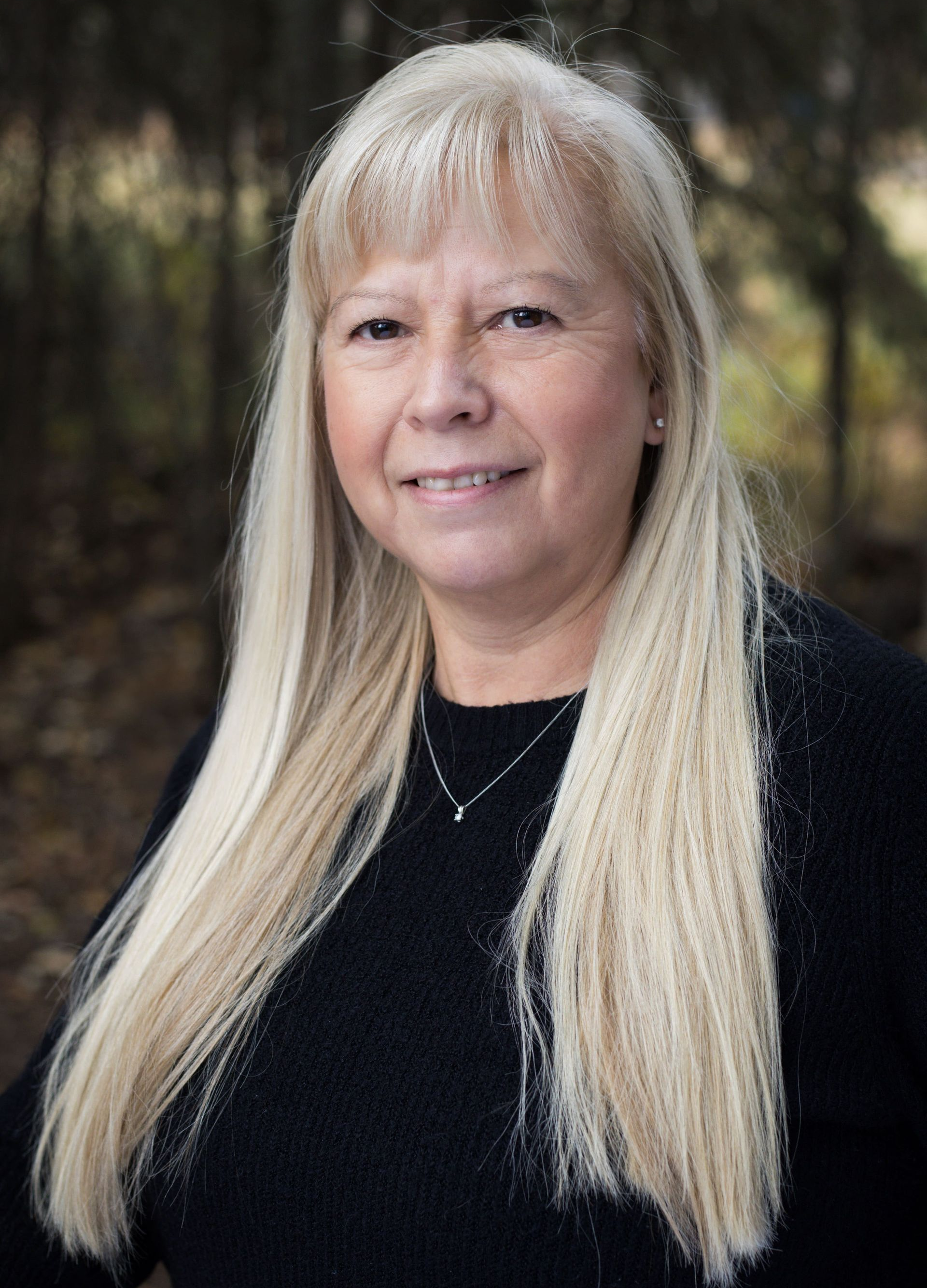 Woman with long blonde hair, smiling, wearing a black sweater, set against a blurred outdoor backdrop.