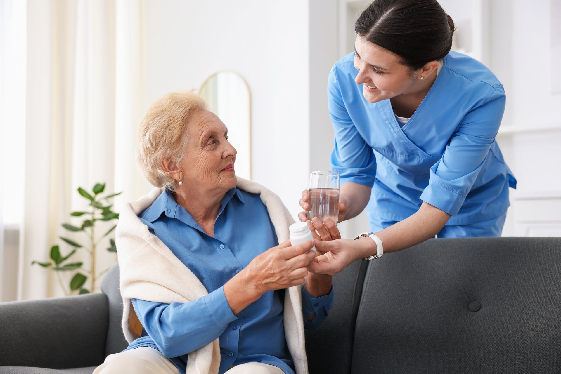 A caregiver in blue scrubs hands a pill and a glass of water to an individual sitting on a gray sofa in a home setting.