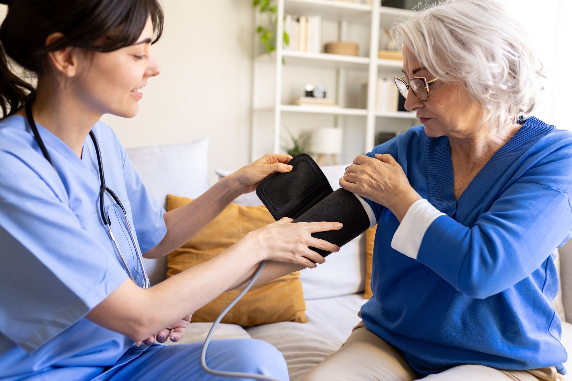 A healthcare worker in blue scrubs helps an individual adjust a blood pressure cuff on their arm in a home setting.
