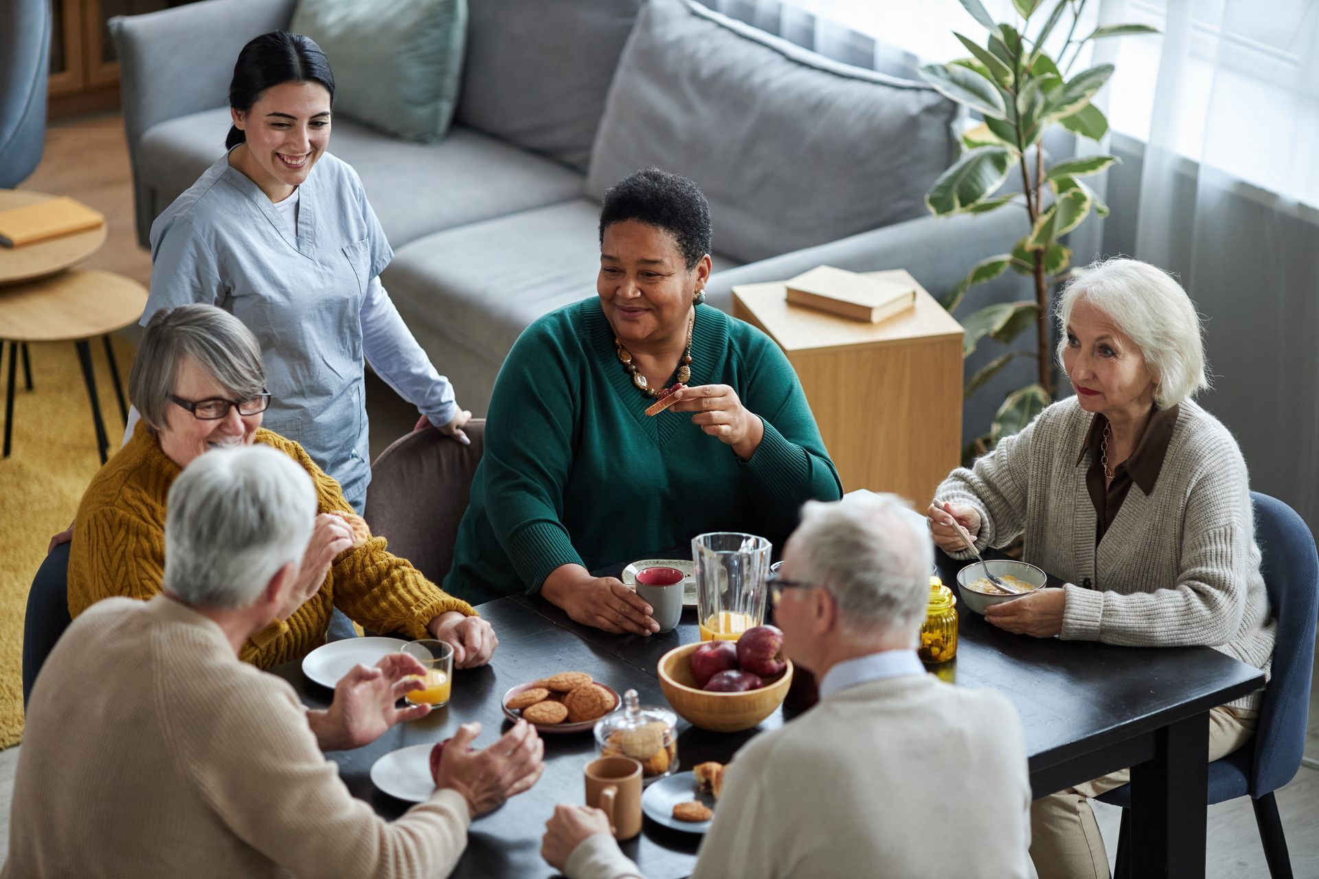 People sit together around a dining table in a bright room, sharing a meal and conversation, with a caregiver standing by.