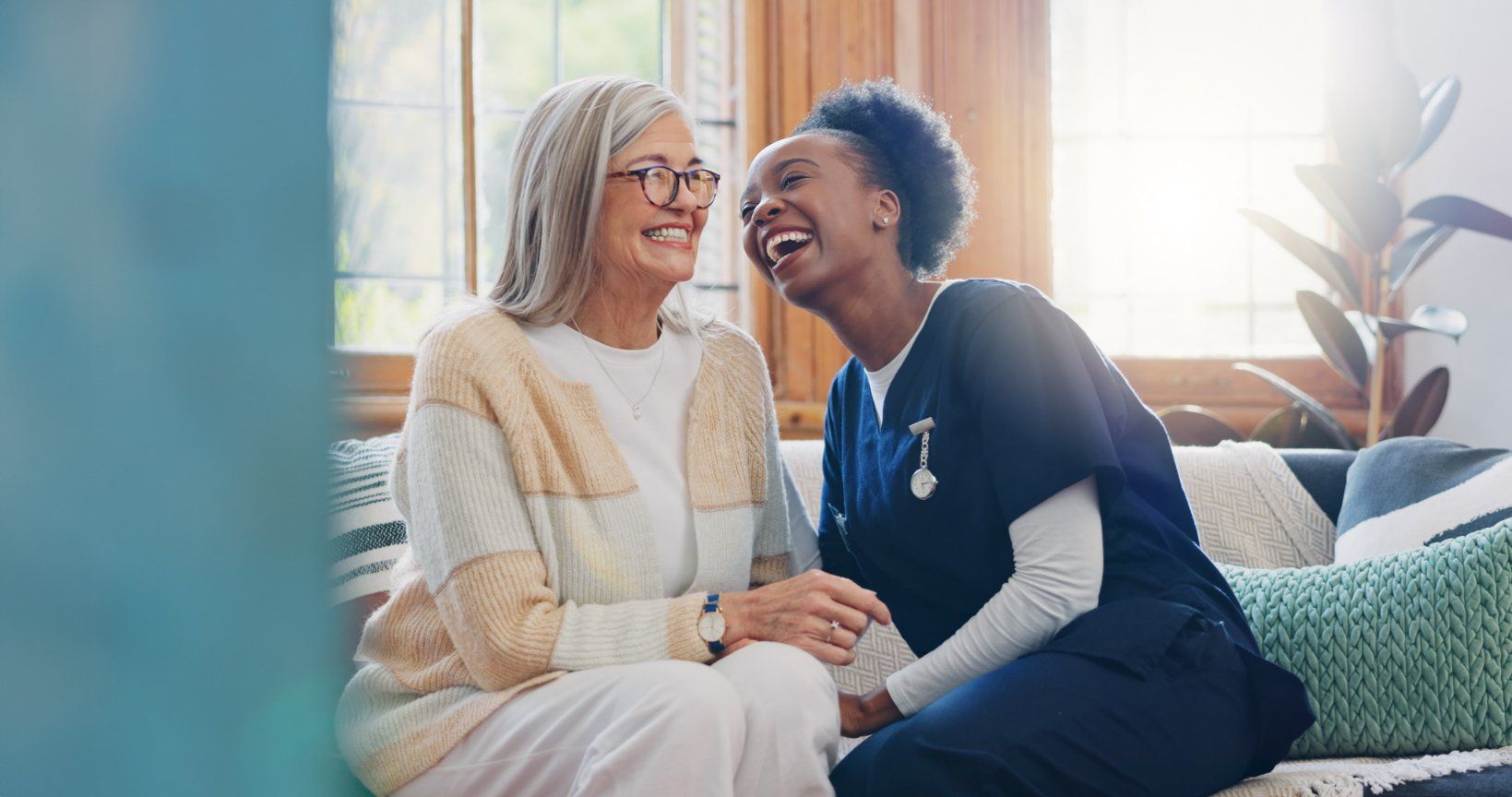 A caregiver and a senior sit together on a sofa, laughing and holding hands in a bright, cozy room.