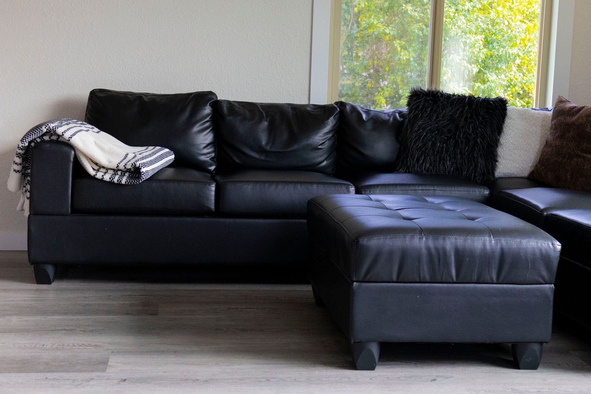 A black leather sectional sofa with a matching ottoman, a throw blanket, and textured decorative pillows in a room.