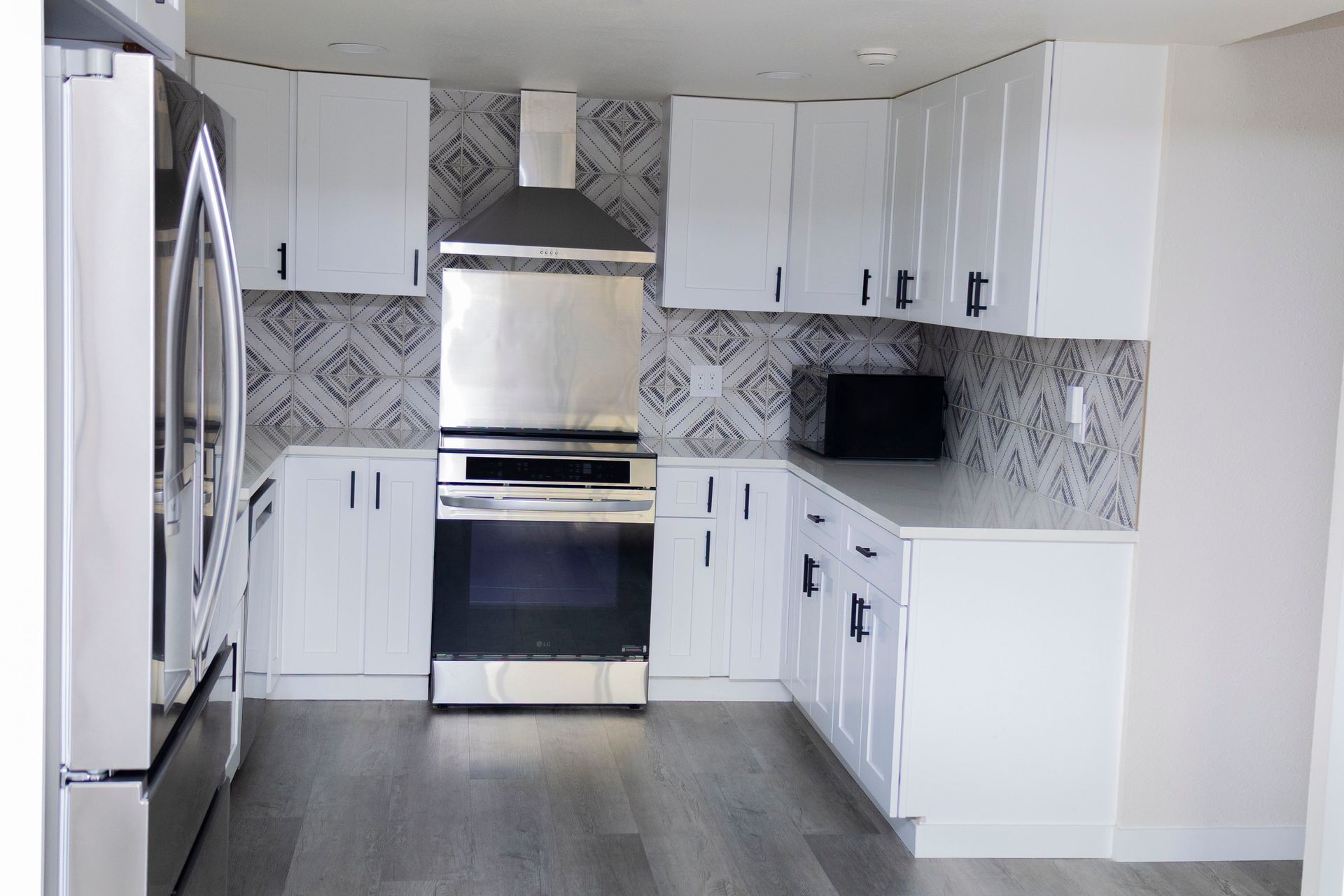 A modern kitchen with white cabinets, stainless steel appliances, and patterned tile backsplash over gray wood flooring.