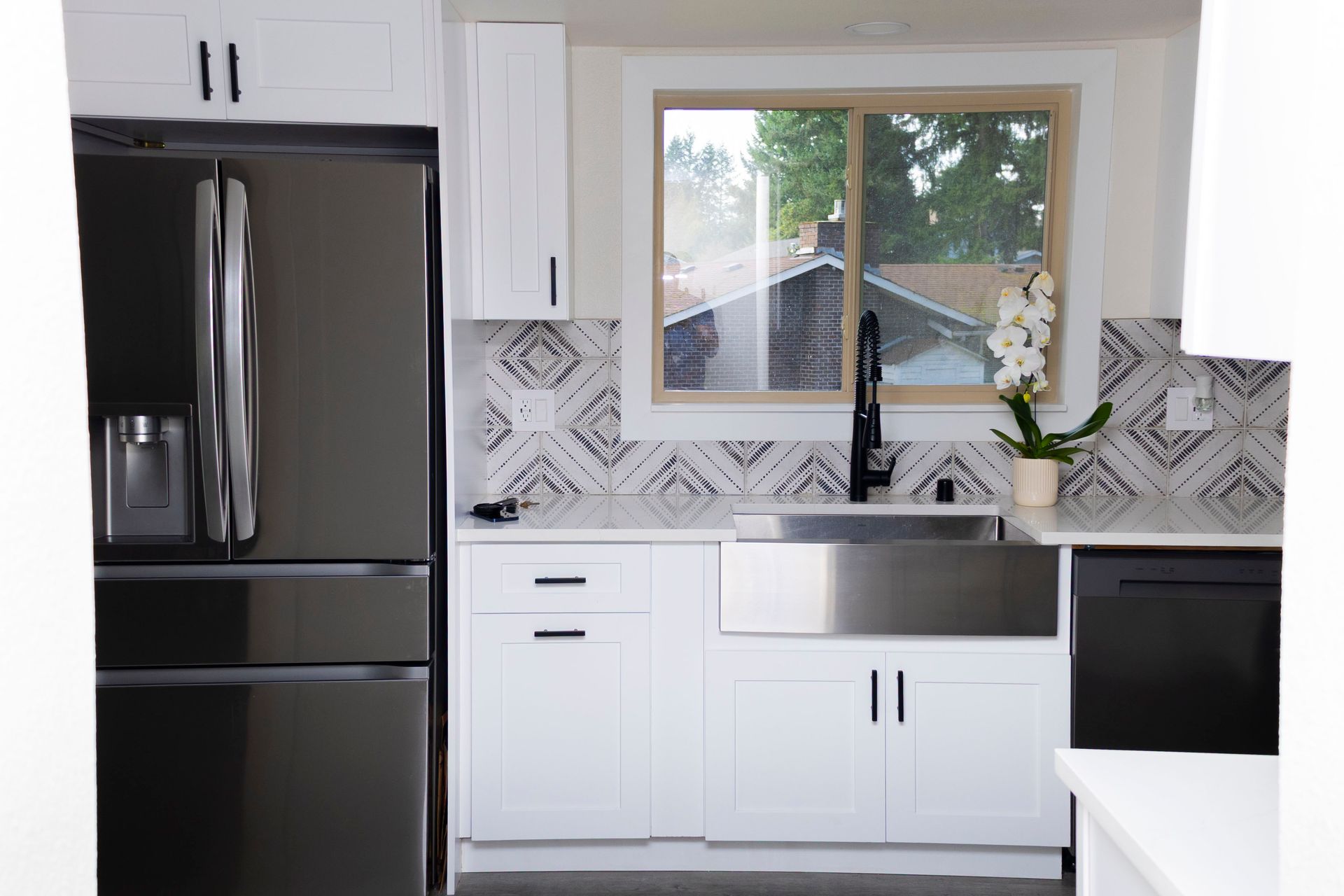 A modern kitchen with a black refrigerator, white cabinets, stainless farmhouse sink, patterned backsplash, and window.