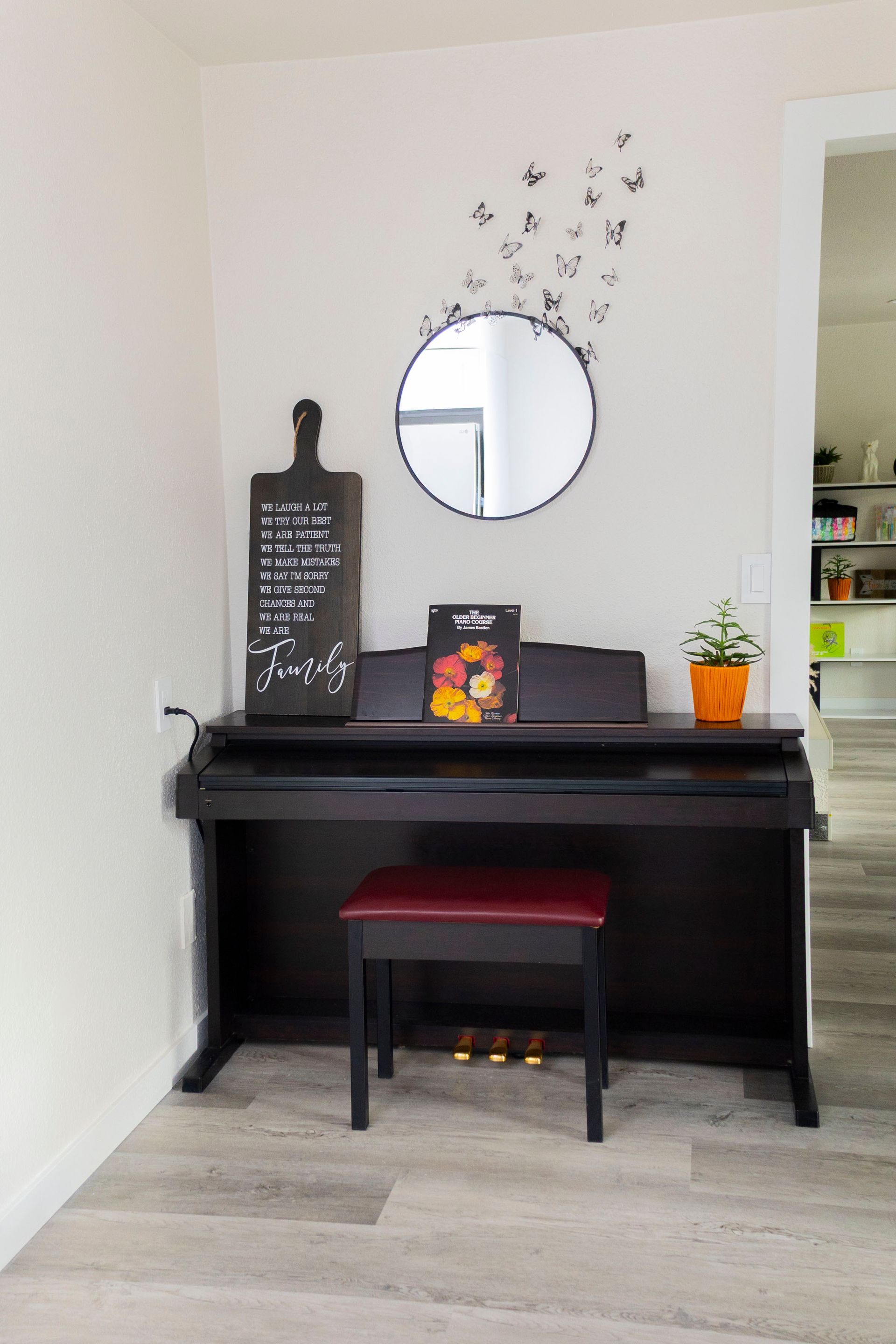 A black digital piano with a red-cushioned bench sits against a white wall below a round mirror and butterfly wall decals.