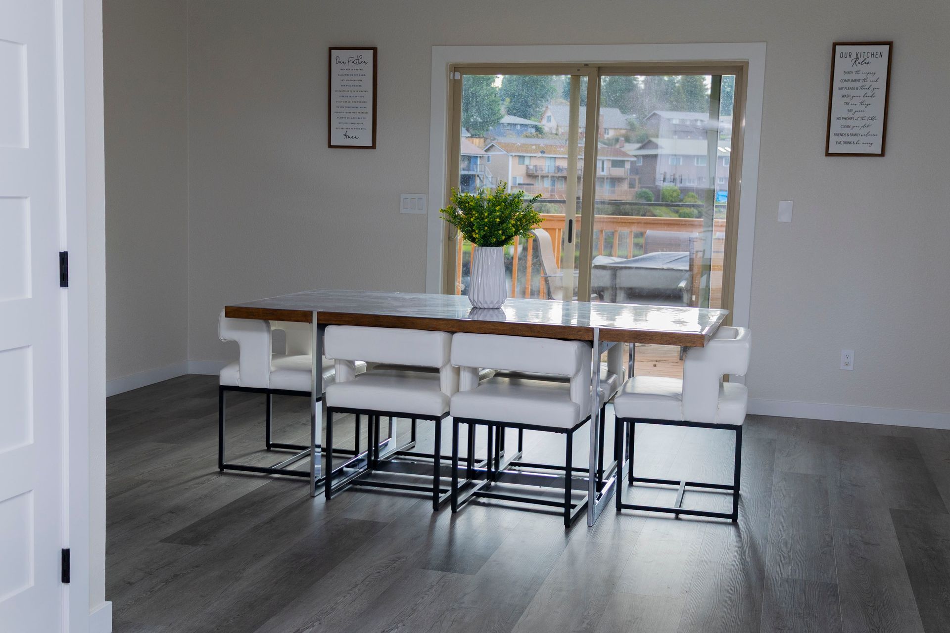 A dining room with a long table, white chairs with black frames, a potted plant centerpiece, and sliding glass doors.