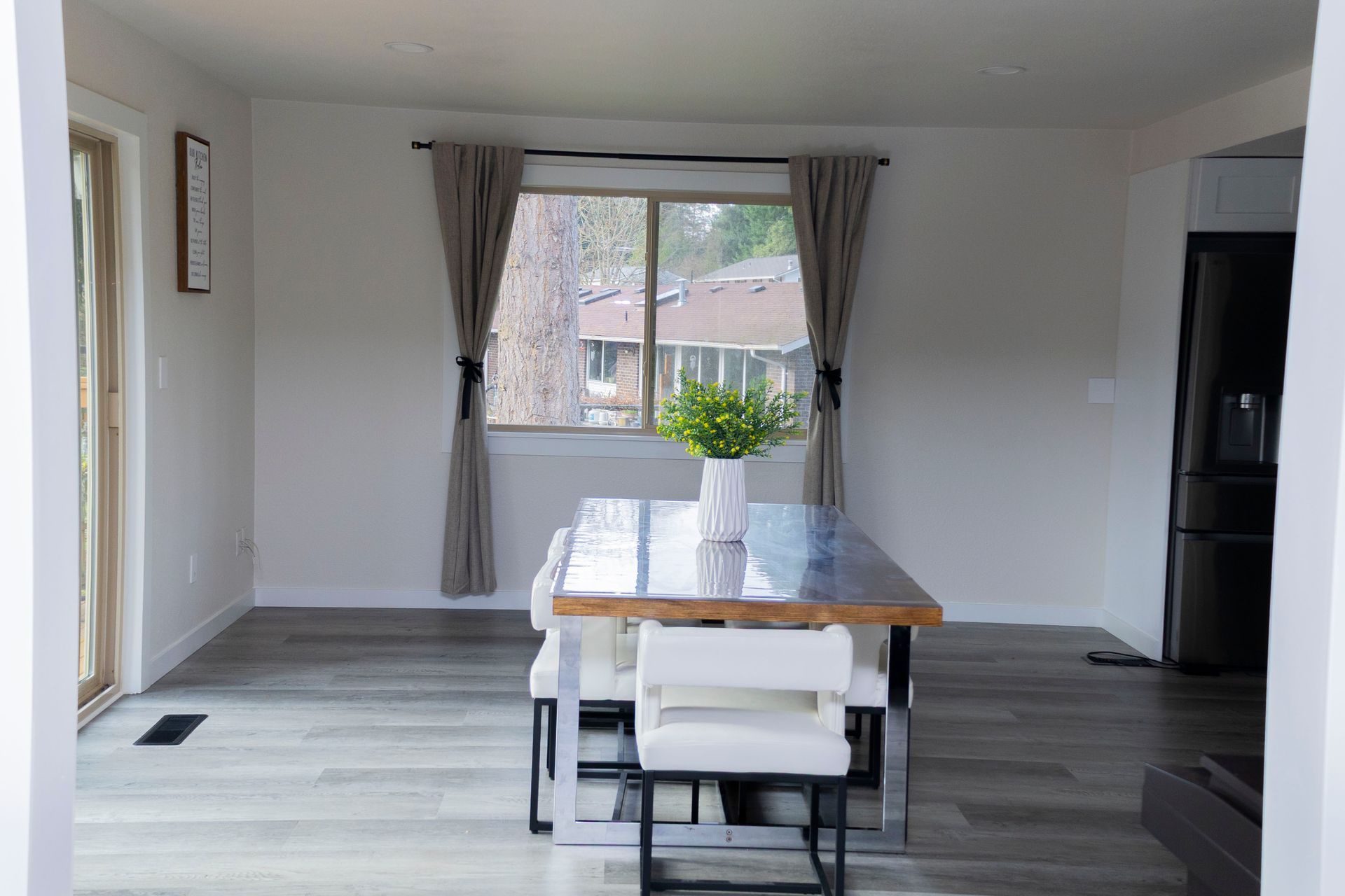 A dining room with light gray wood floors, a wooden table with white chairs, and a window with beige curtains.
