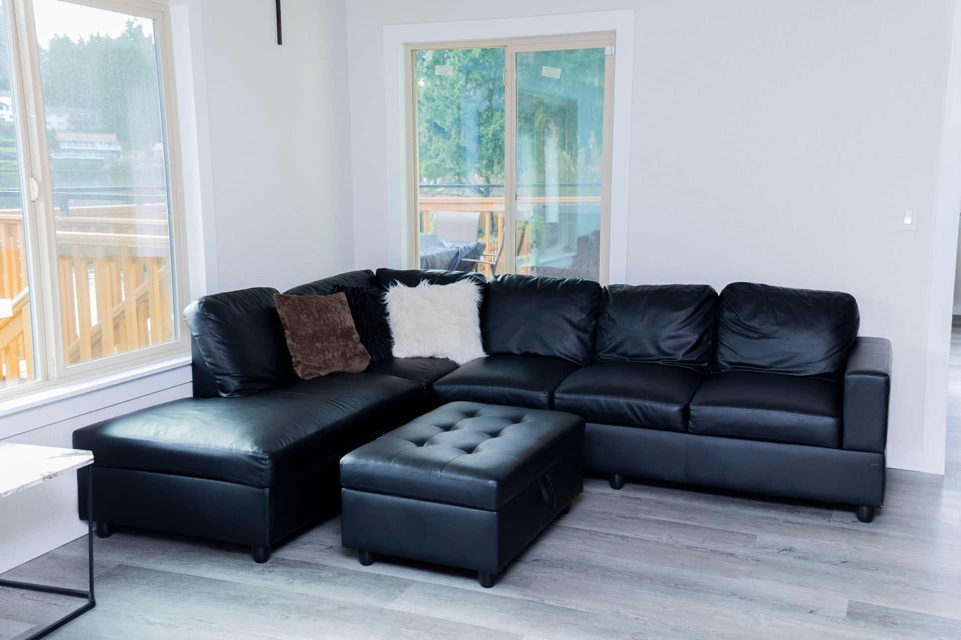 A black leather sectional sofa with a matching storage ottoman in a modern room with large windows and light wood floors.