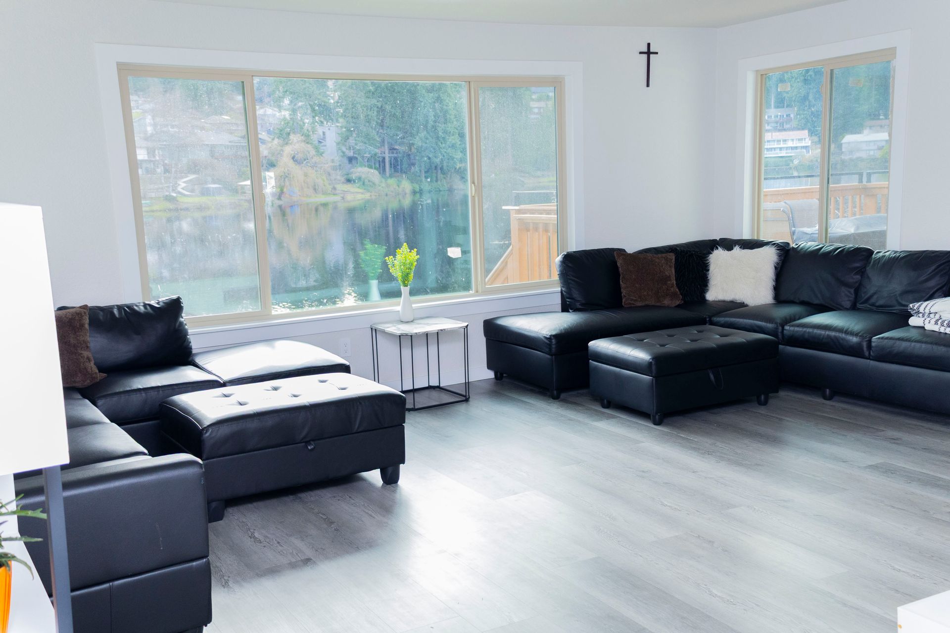 A spacious, modern living room featuring two black sectional sofas, a matching ottoman, and large windows with a lake view.