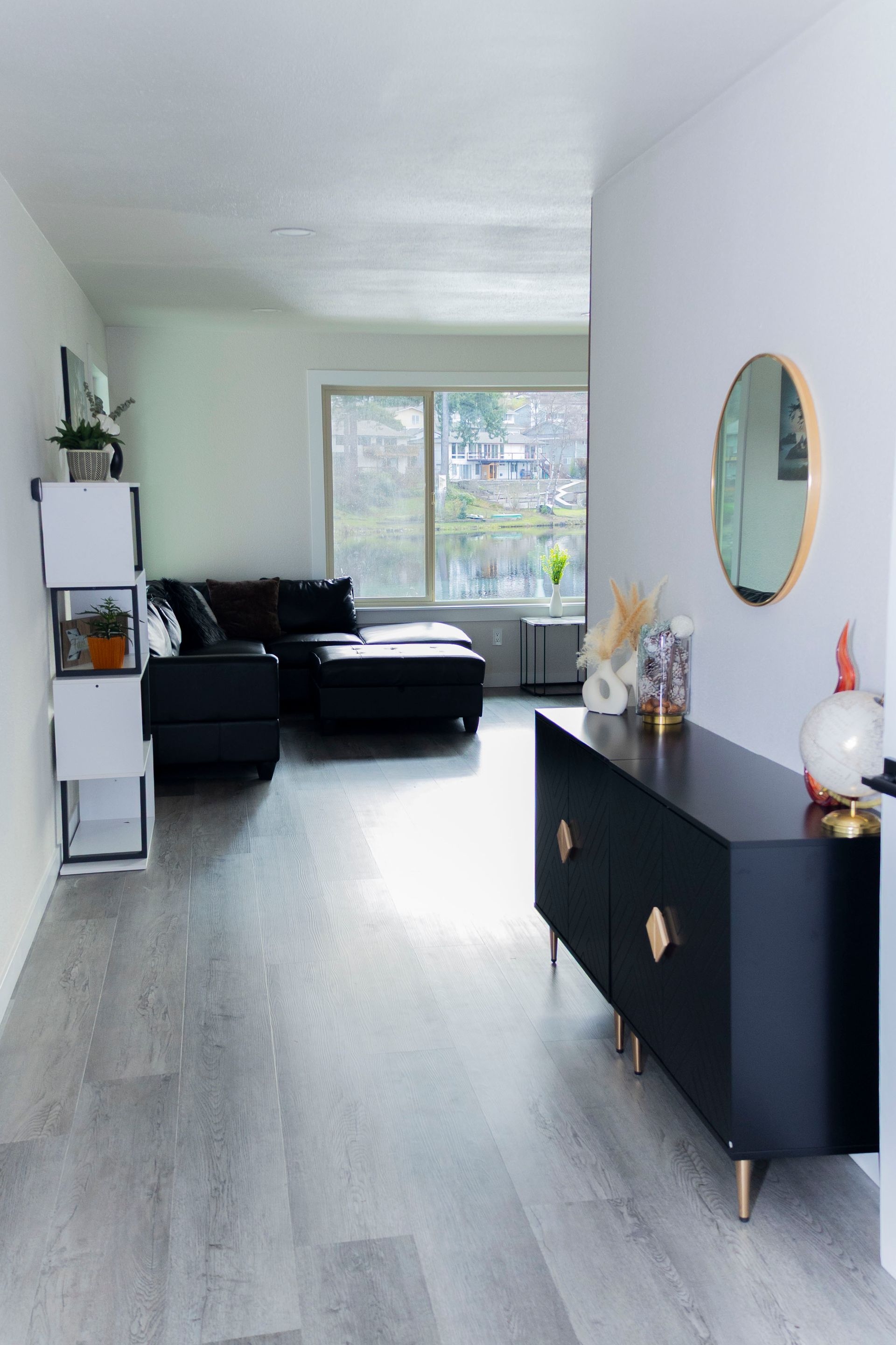 A modern living room with a black sideboard, round mirror, and white shelves, leading to a couch by a large window.