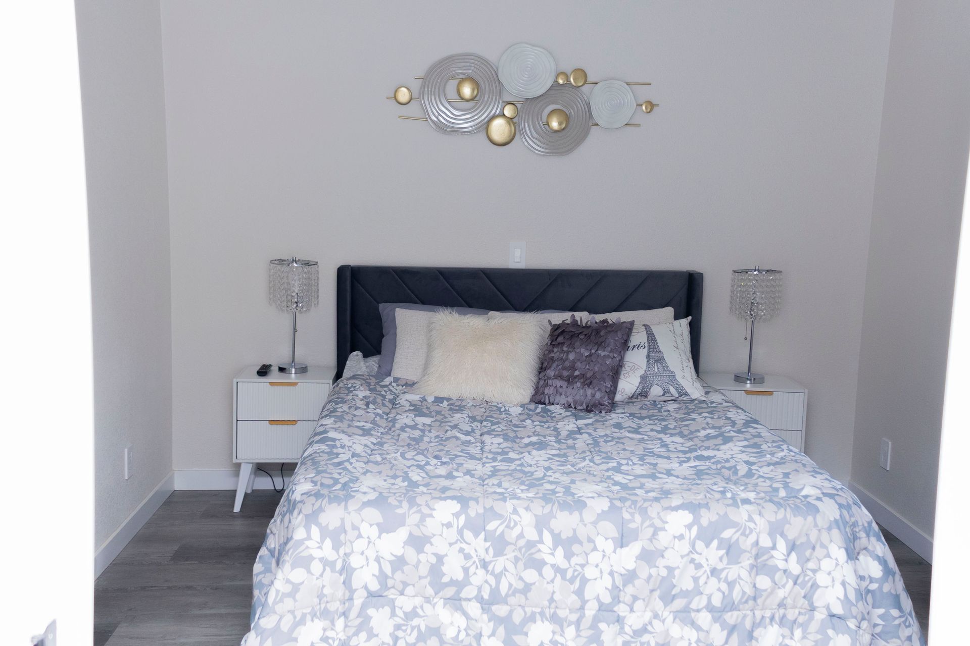 A bedroom featuring a dark headboard, a white floral patterned bedspread, two nightstands with lamps, and wall art.