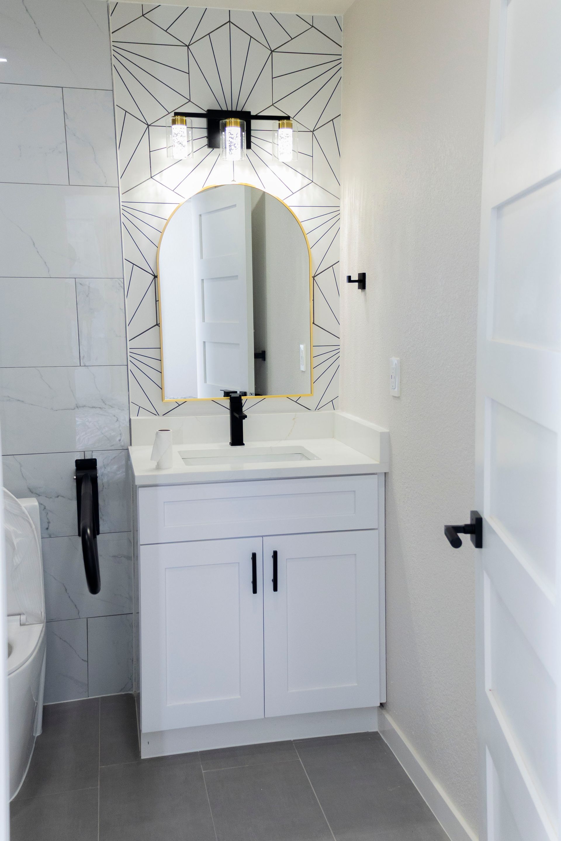 A modern bathroom vanity with a white cabinet, black faucet, arched mirror, and geometric patterned accent wallpaper.