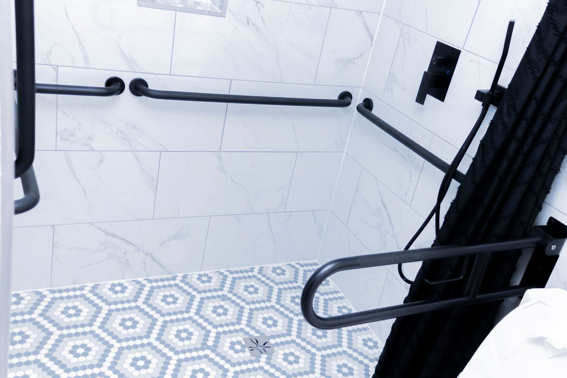 An accessible shower with white marble-style walls, patterned tile floors, and multiple black support grab bars.