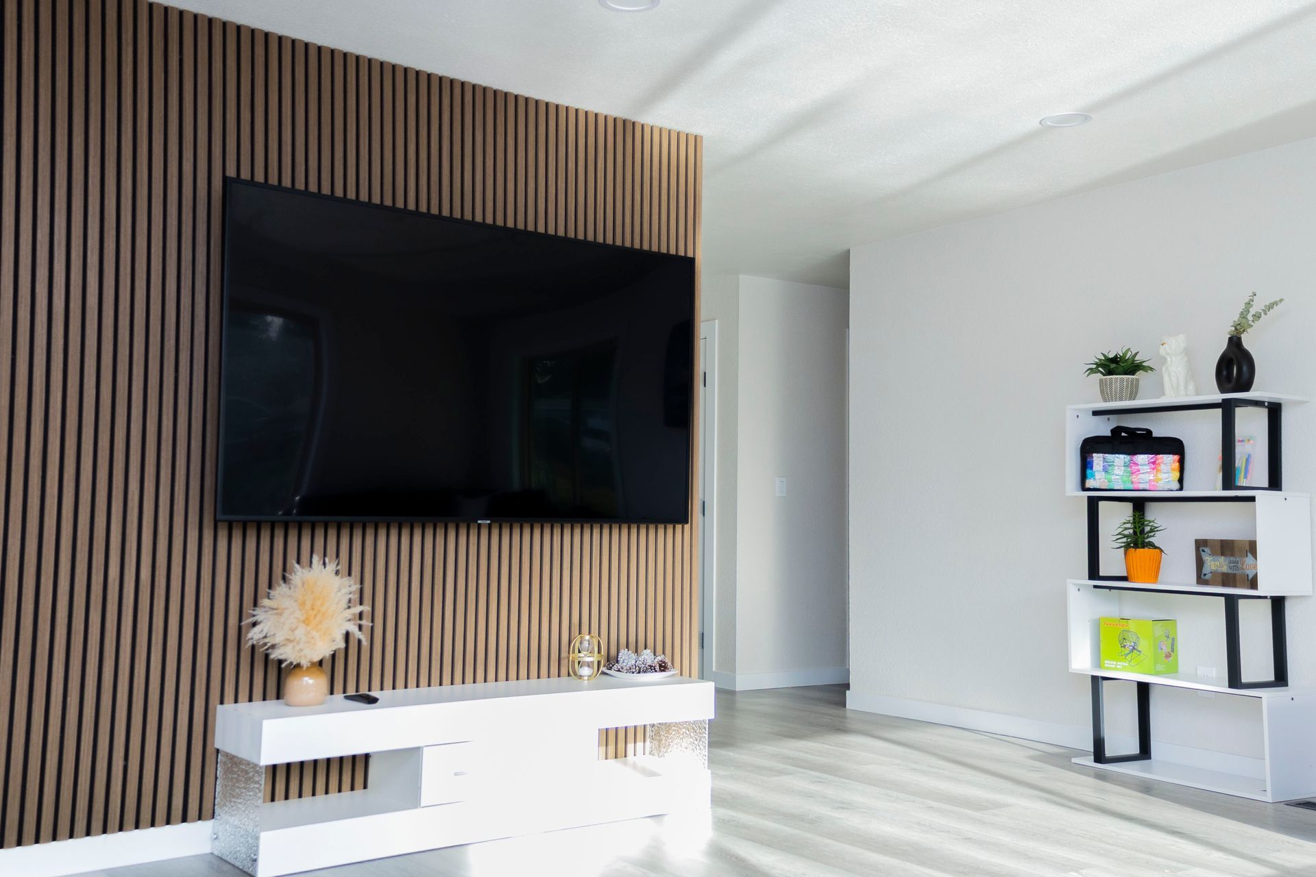 A wall-mounted TV over a white media console against a wood-slat accent wall, with an adjacent modern white bookshelf.