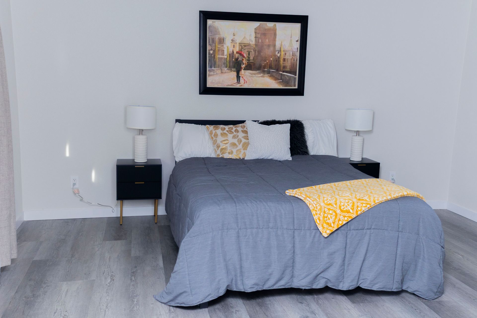 A grey bed with a yellow throw blanket, flanked by two black nightstands with lamps, and a framed wall painting above.