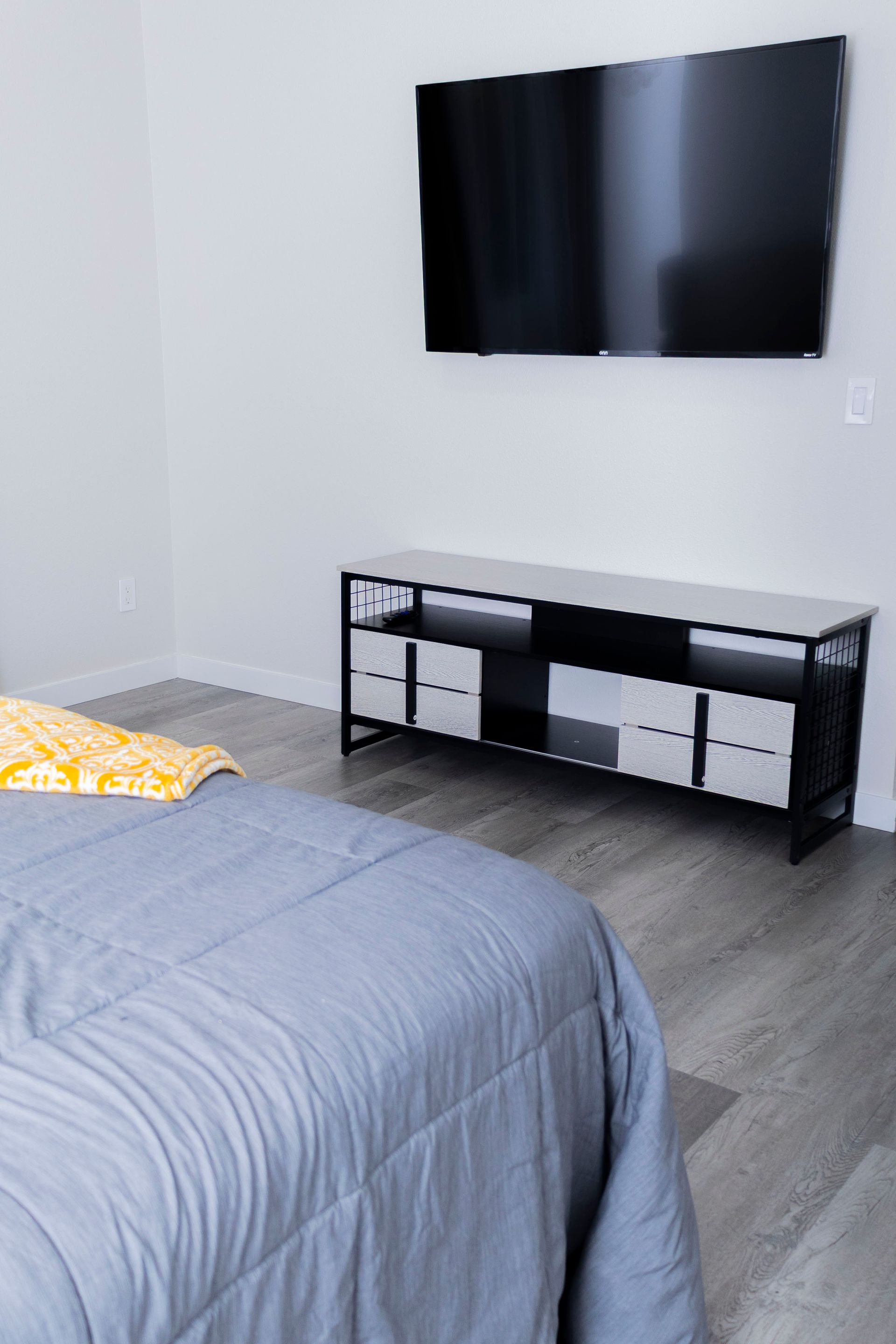 A wall-mounted TV sits above a black, multi-compartment storage console in a minimalist bedroom with grey flooring.