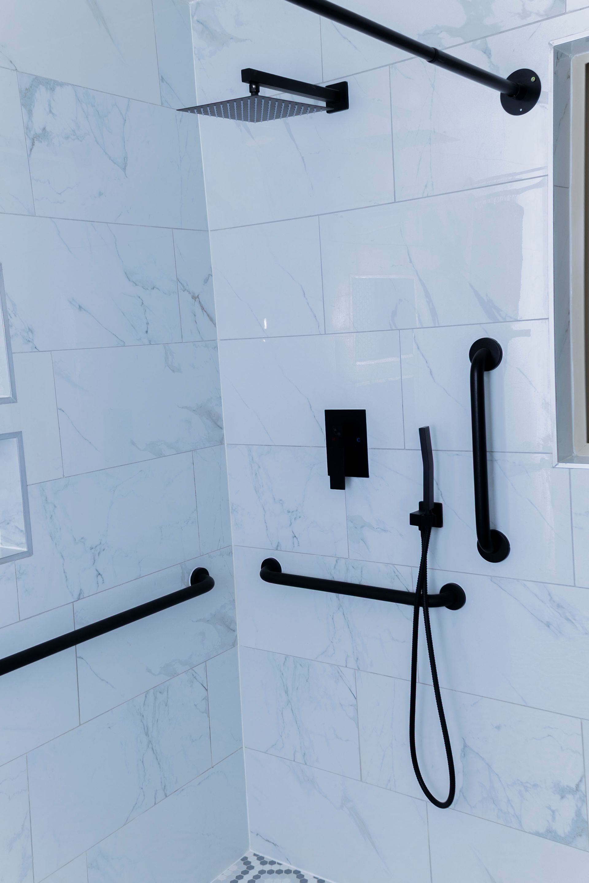 A modern shower with white marble-style tiles, matte black fixtures, a rain shower head, handheld sprayer, and grab bars.