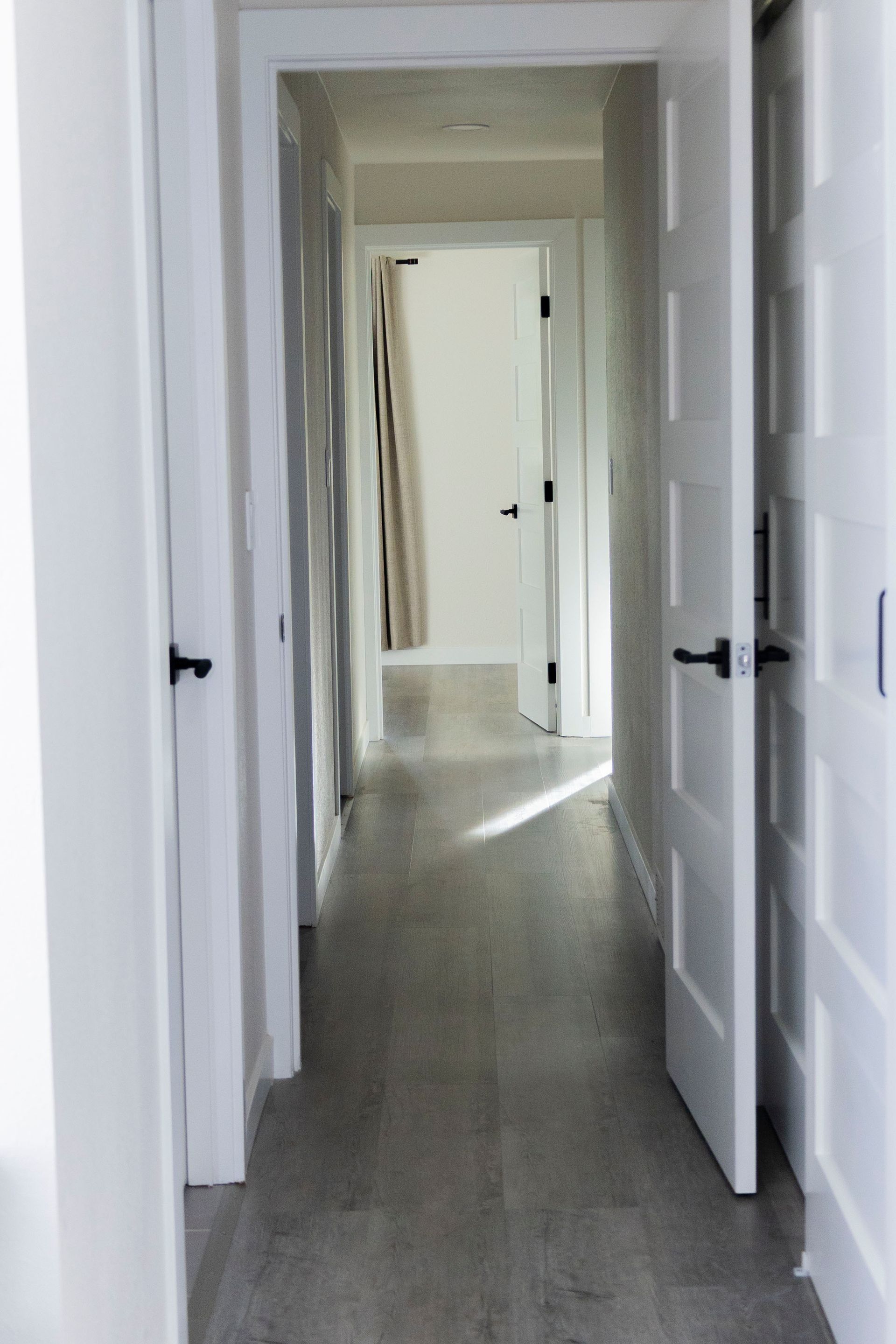 A hallway with light gray wood flooring, white walls, and white doors leading to another room with a draped window.