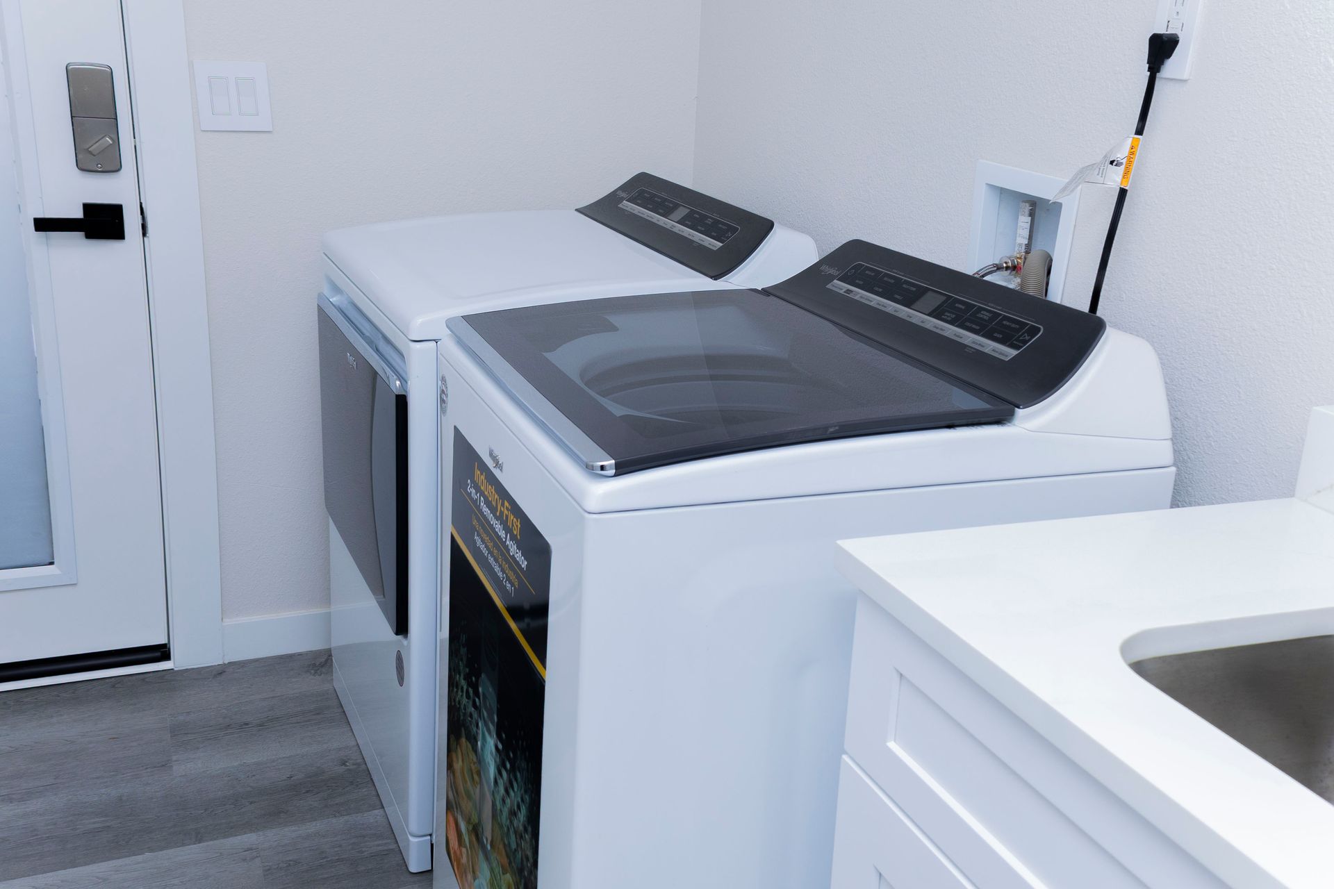 A pair of white Whirlpool washing machines sit in a laundry room next to a white sink and a door with a keyless lock.