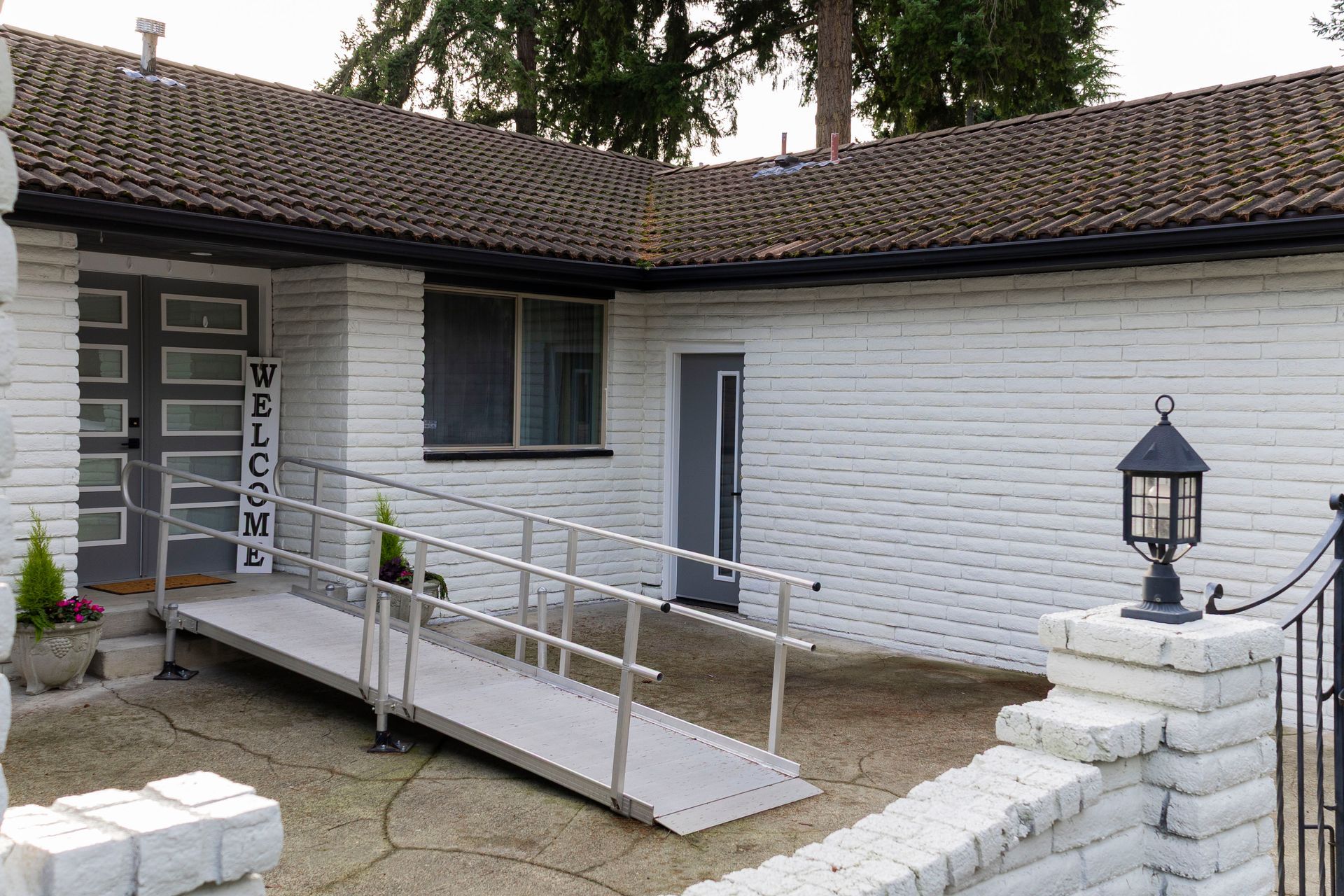 A white brick home entrance featuring a metal wheelchair ramp leading to a set of grey double doors.