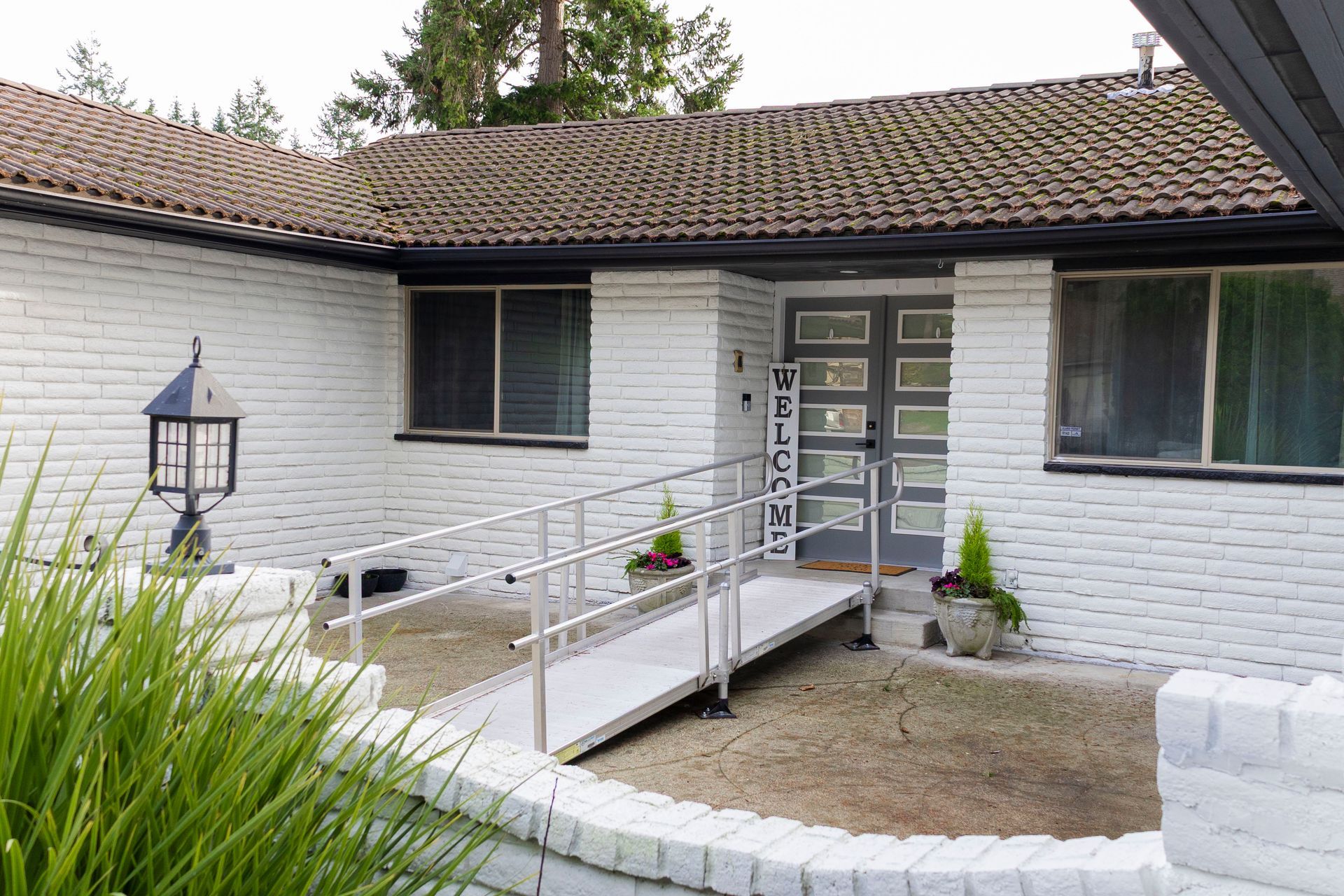 A white brick house with a tile roof and a metal wheelchair ramp leading to a gray double front door.