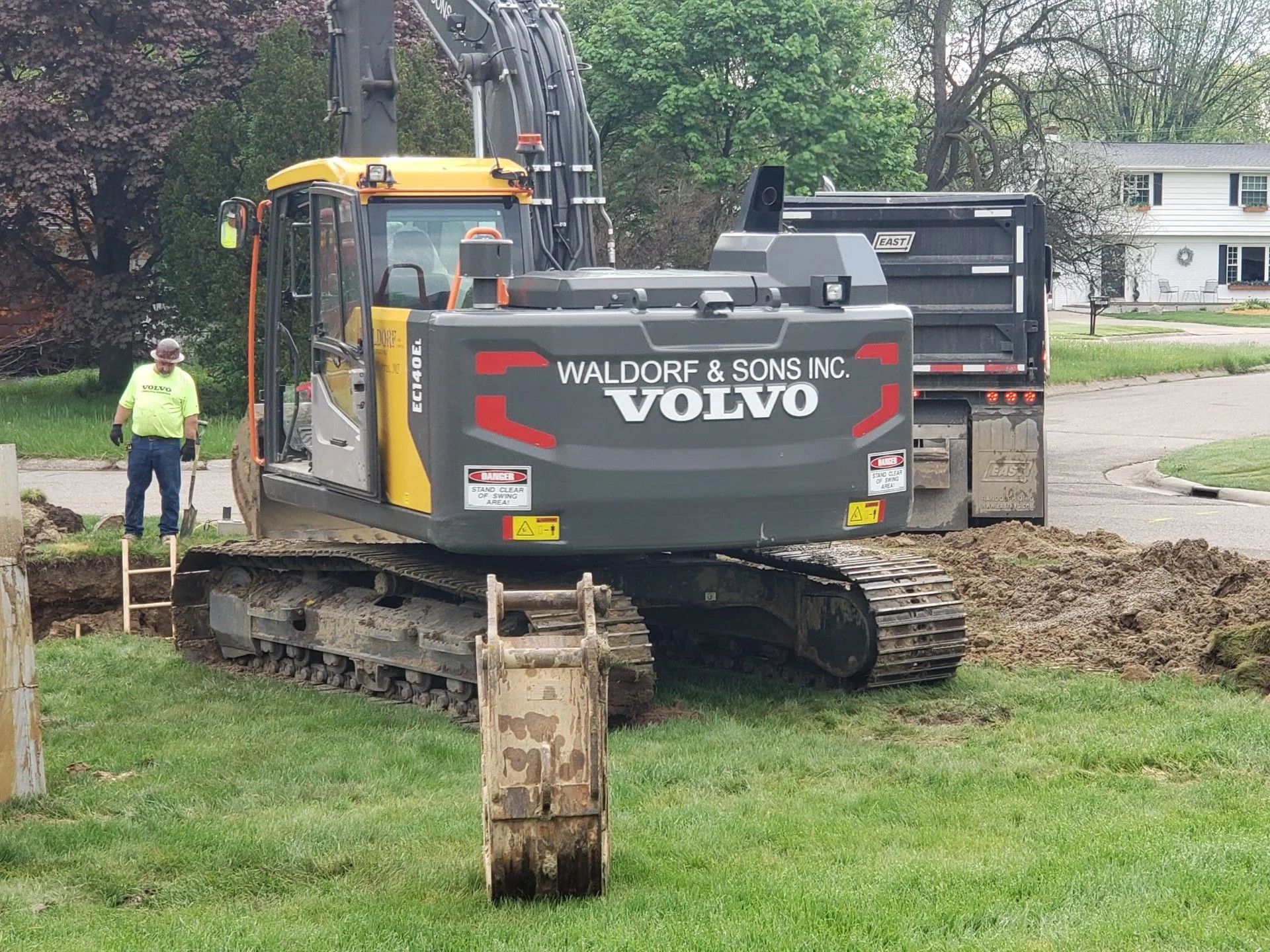 Excavator digging near a dump truck; a worker observes. Outdoors, on grass, daytime. Volvo machinery, grey and yellow.