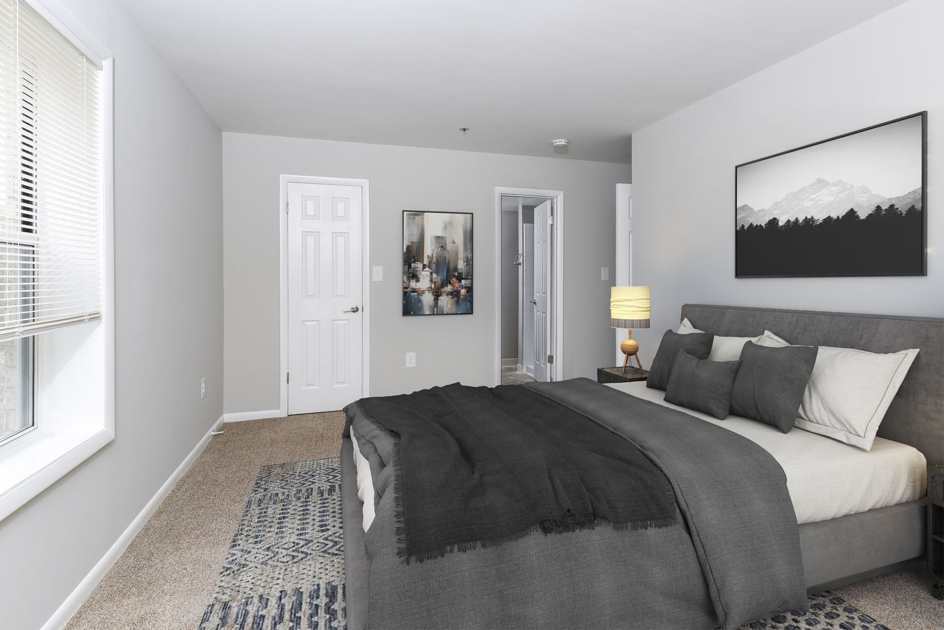 Bedroom with gray bed, pillows, nightstand lamp, and window blinds.