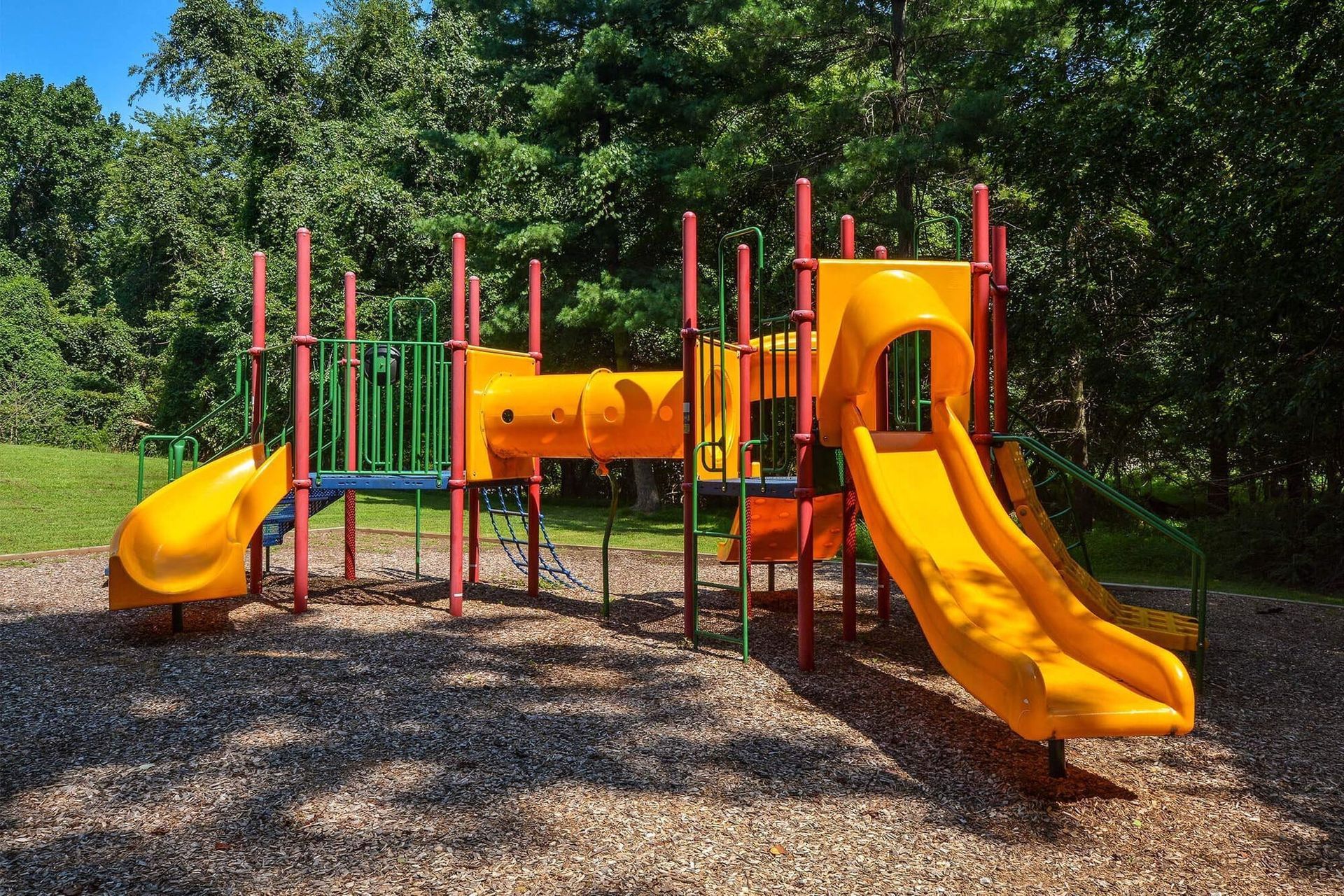 Playground with bright yellow slides and red-green climbing frames in a leafy outdoor area.