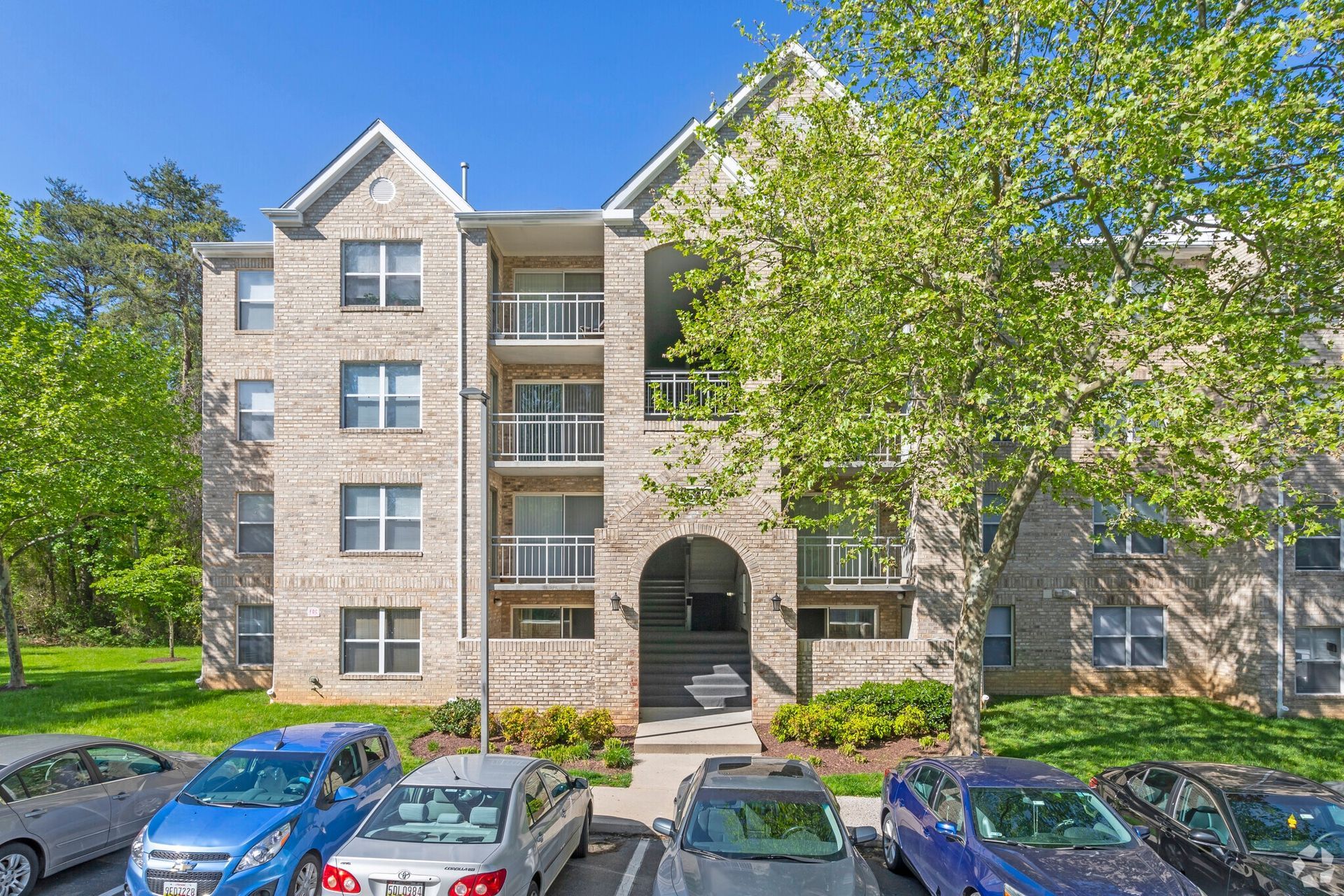 Exterior view of a brick apartment building with balconies, trees, and parked cars.